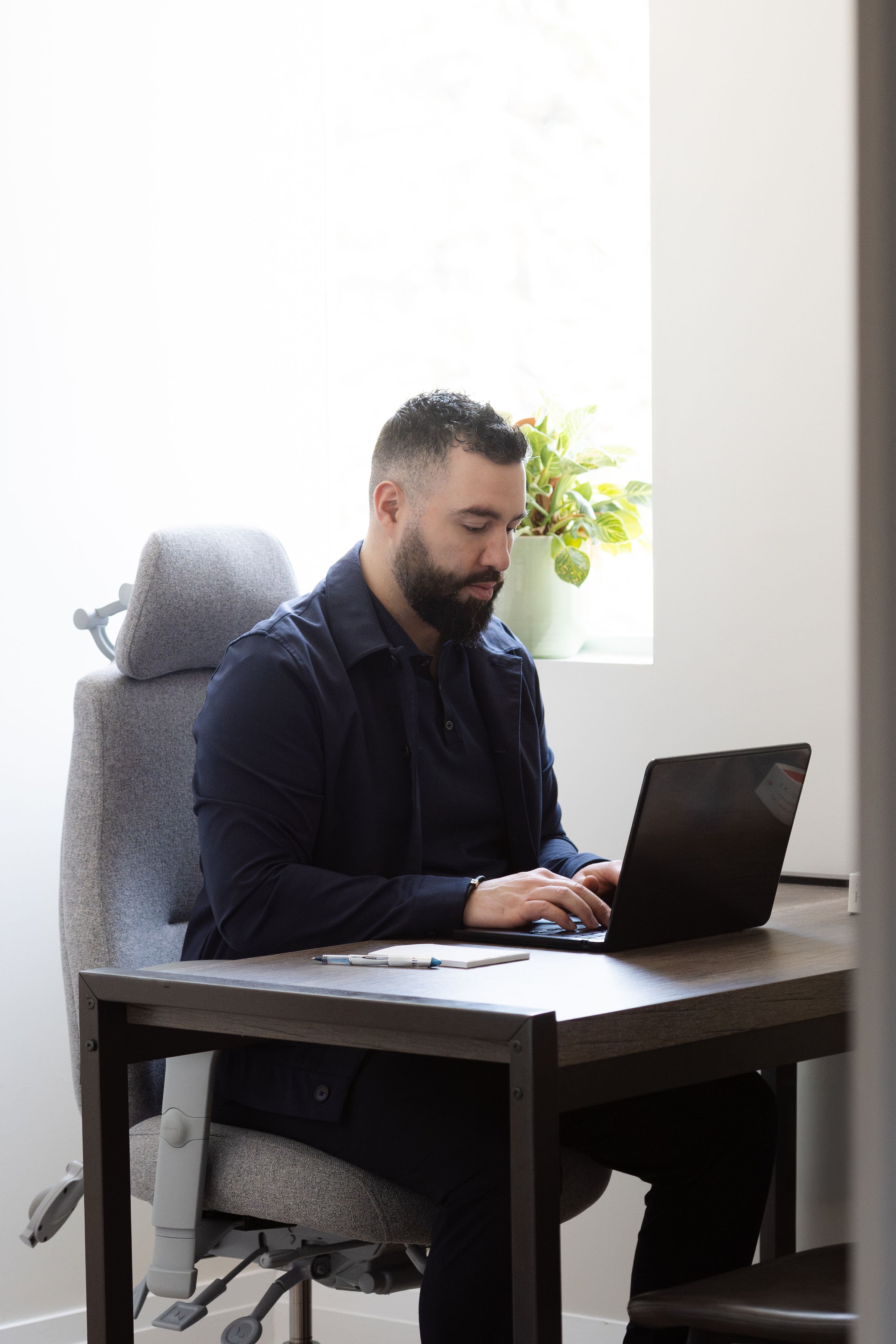 A man with short dark hair and a beard, wearing a dark blue shirt, sitting at a desk in front of a laptop with a closed expression, in an office with a light-colored wall, a window, and a potted plant in the background.