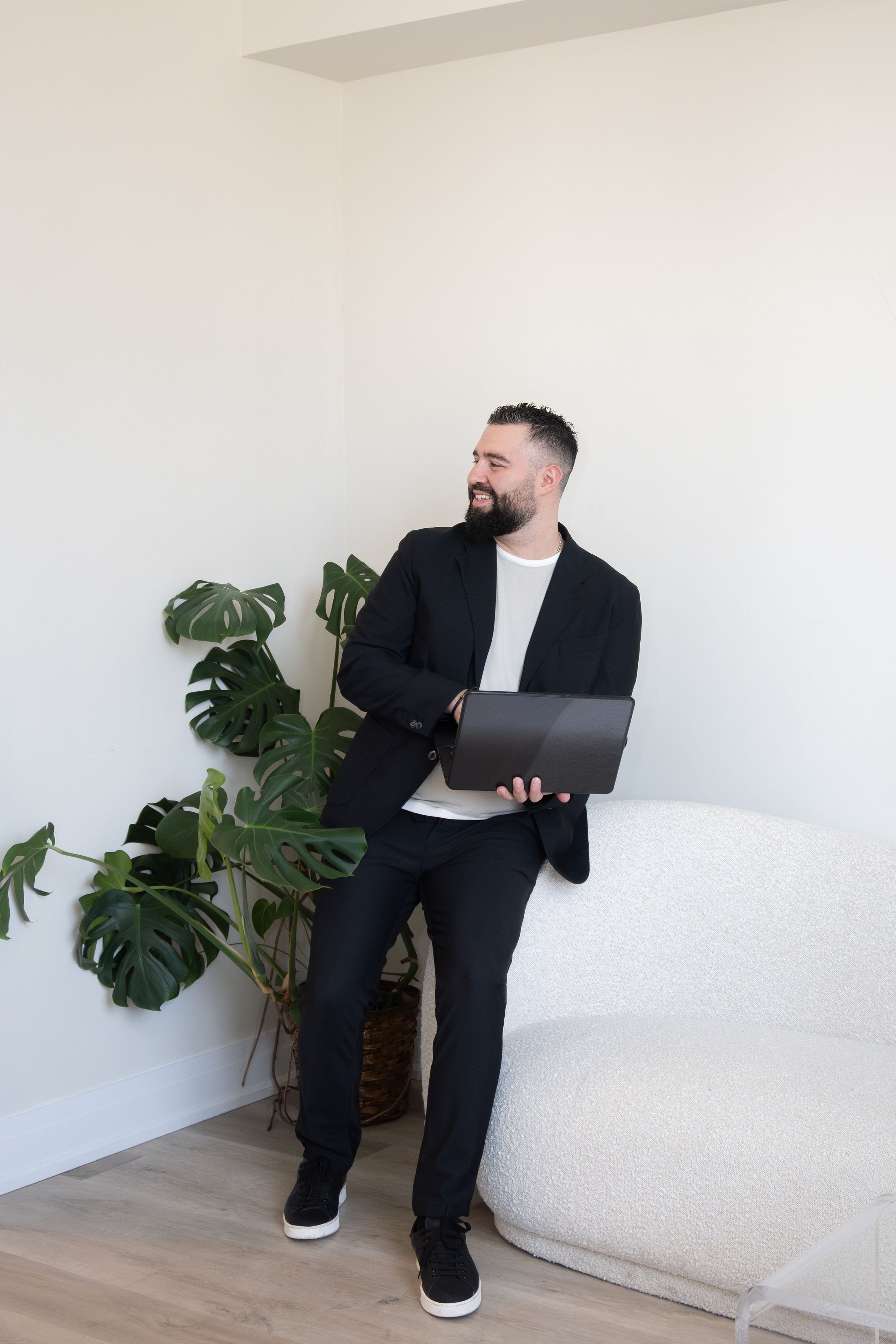 A man with a beard, dressed in a black blazer, white shirt, and black pants, standing in a minimalistic room with a white wall, holding a tablet. There is a large potted monstera plant to his left and a white upholstered chair to his right.