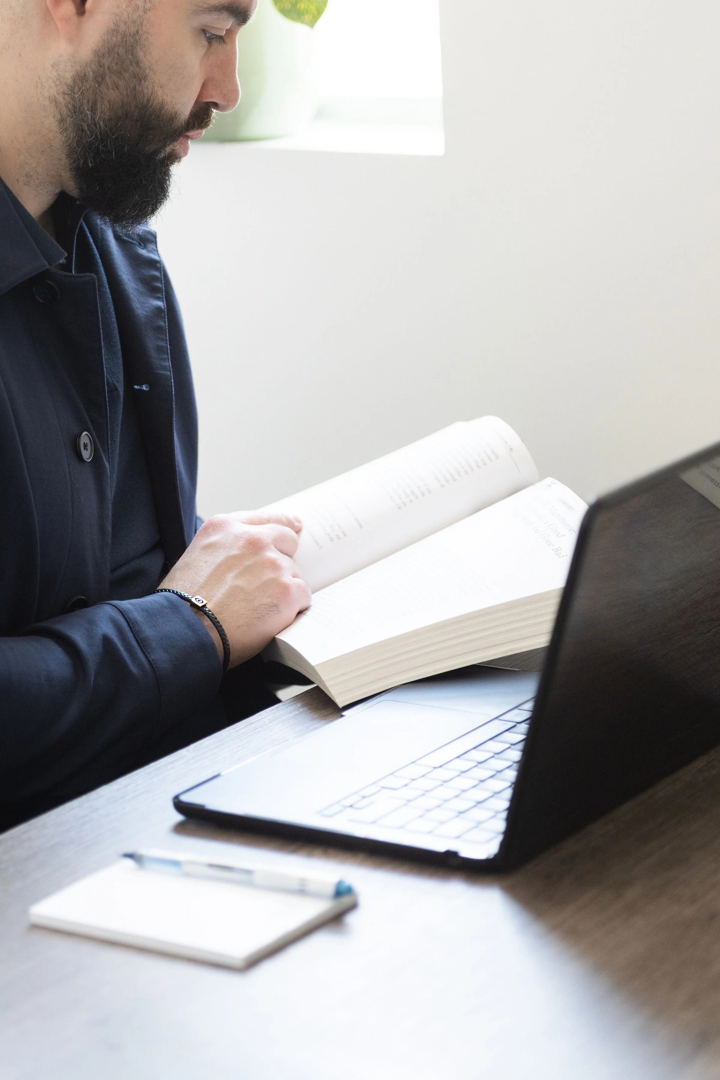 A man reading a book at a desk with a laptop and notebook in front of him.