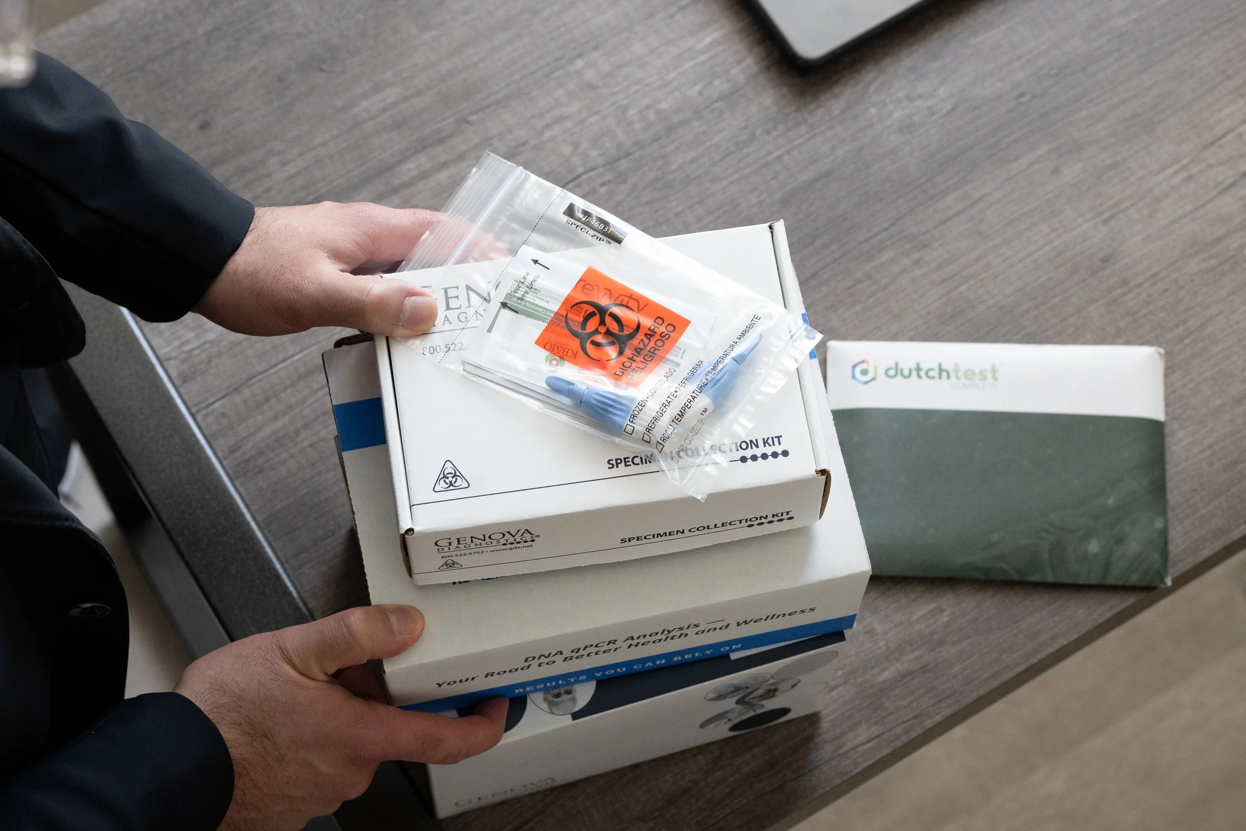 Person holding a medical specimen collection kit box on a table, with biohazard warning and testing supplies inside the kit, and a box labeled 'Dutch Test' nearby.
