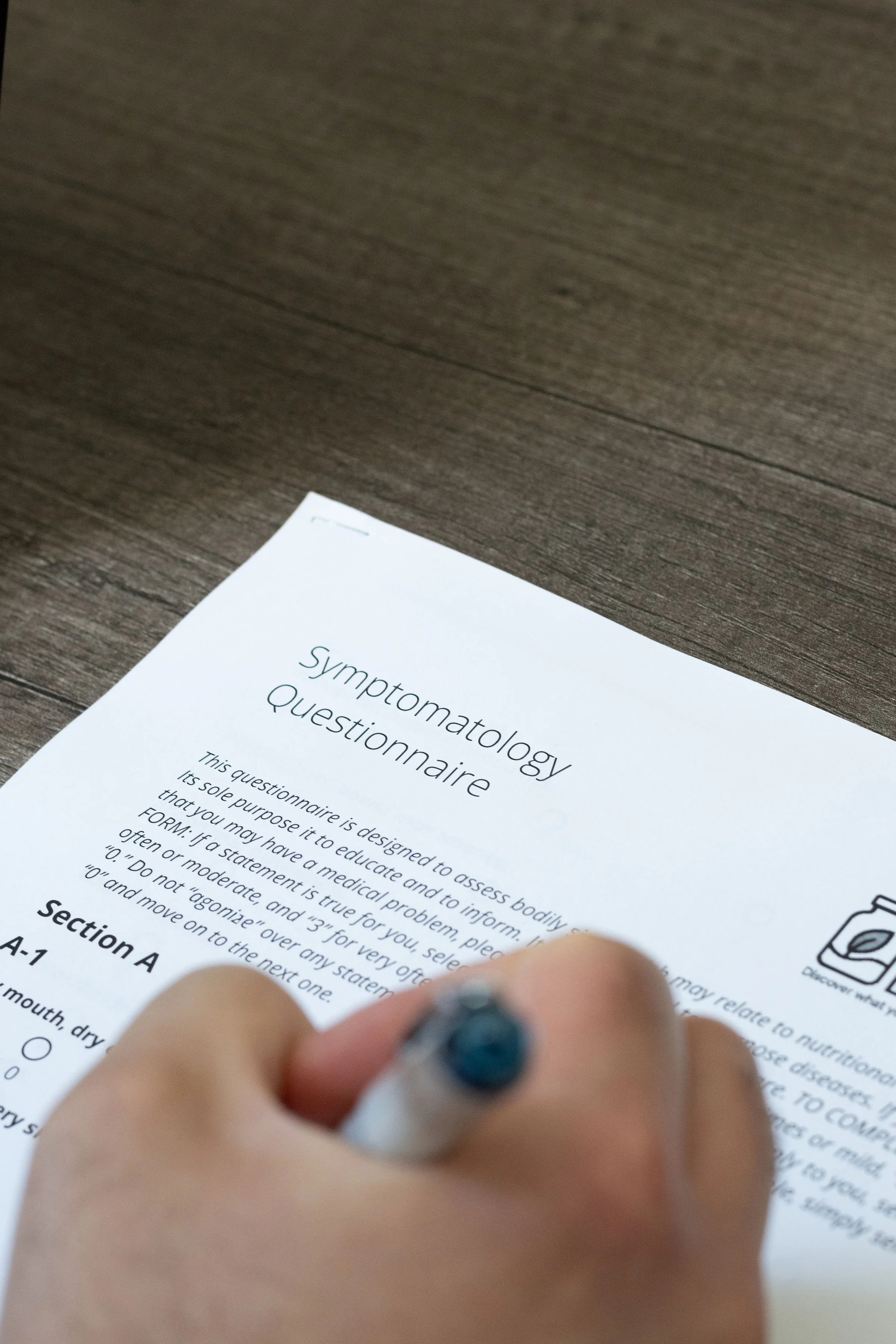A person with painted nails holding a pen, filling out a symptomology questionnaire on a wooden table.