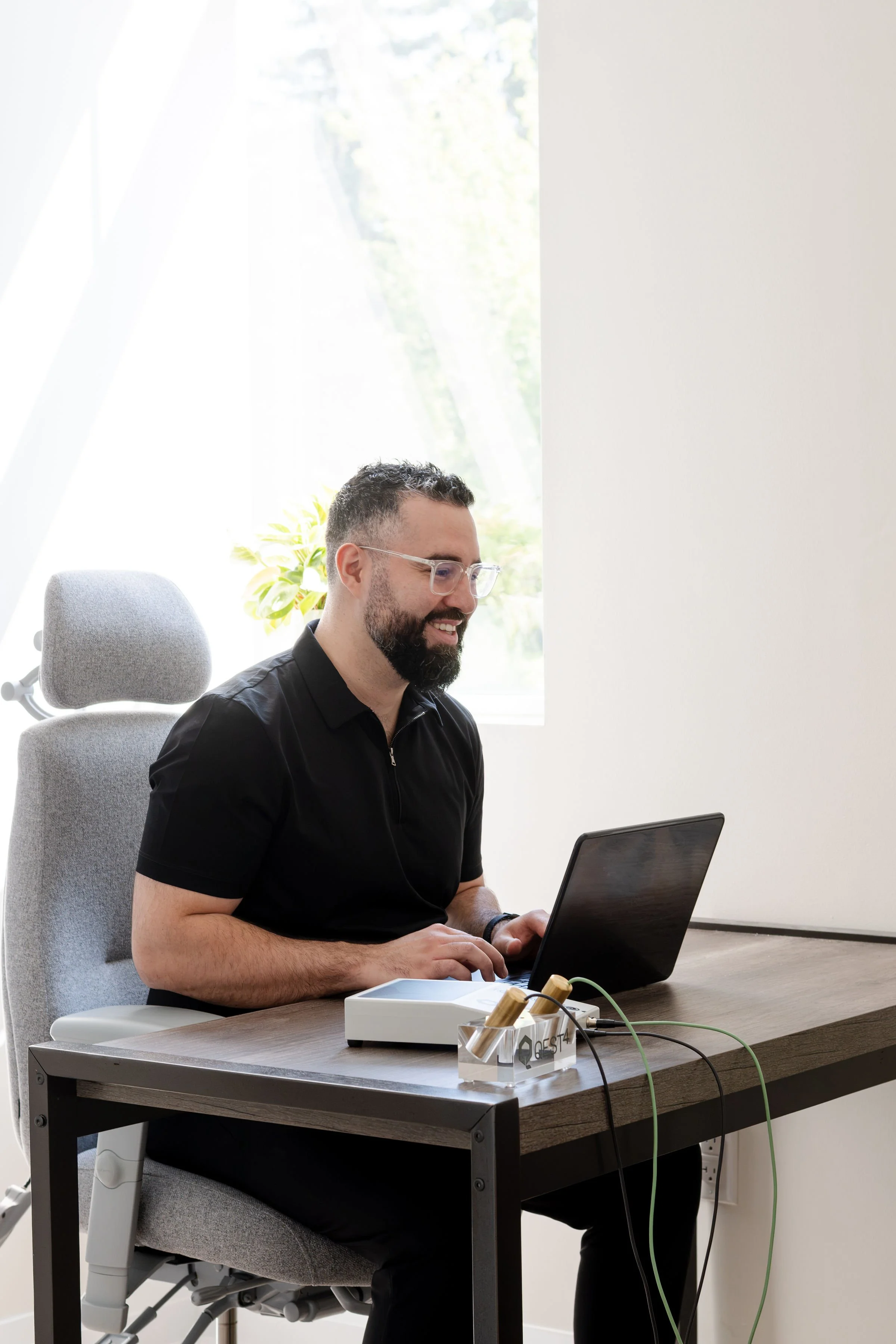 A man with glasses and a beard sitting at a desk, using a laptop, in a well-lit modern office with a plant and window in the background.