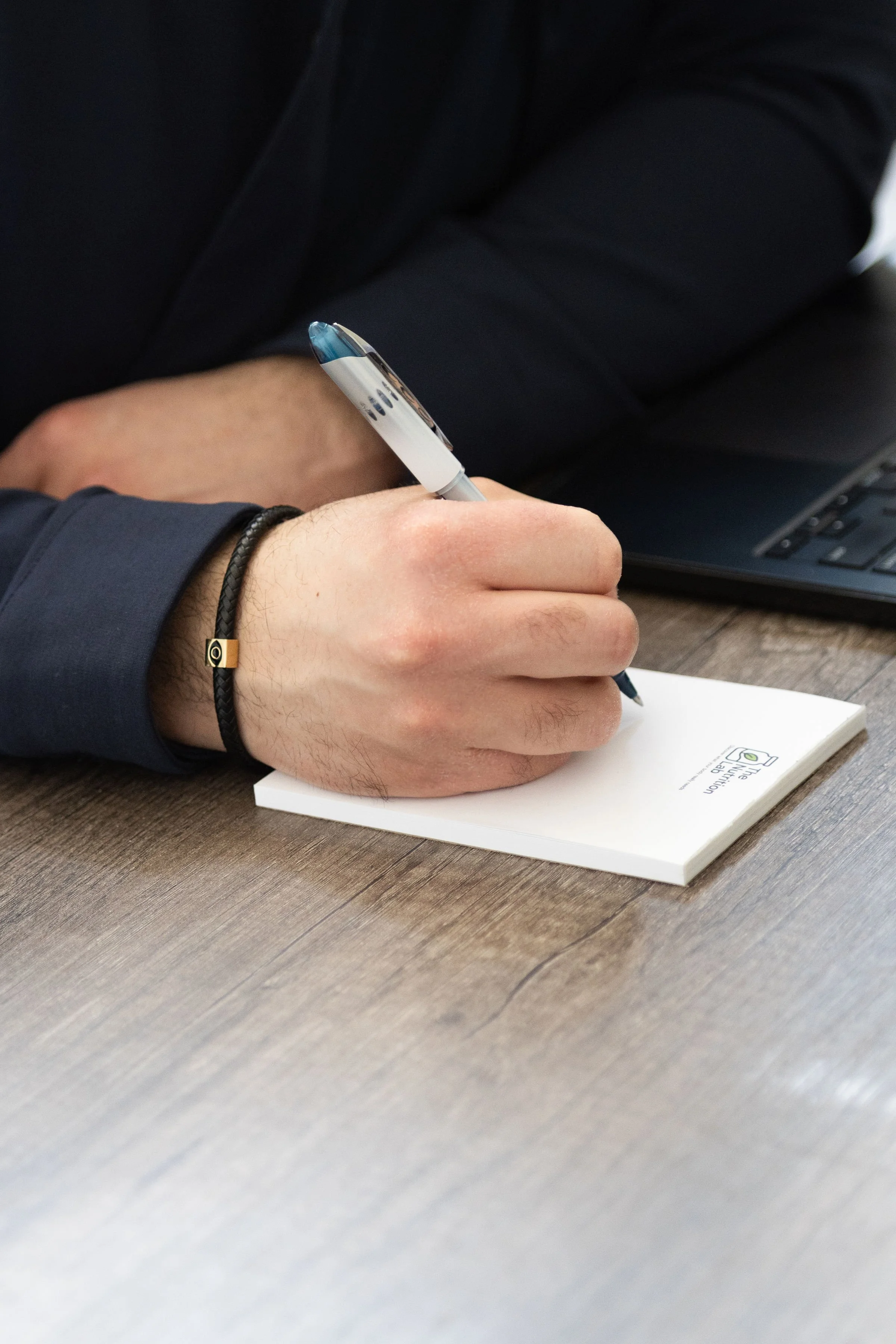 Person writing with a pen on a notepad at a desk, wearing a dark long-sleeved shirt, with a laptop nearby.