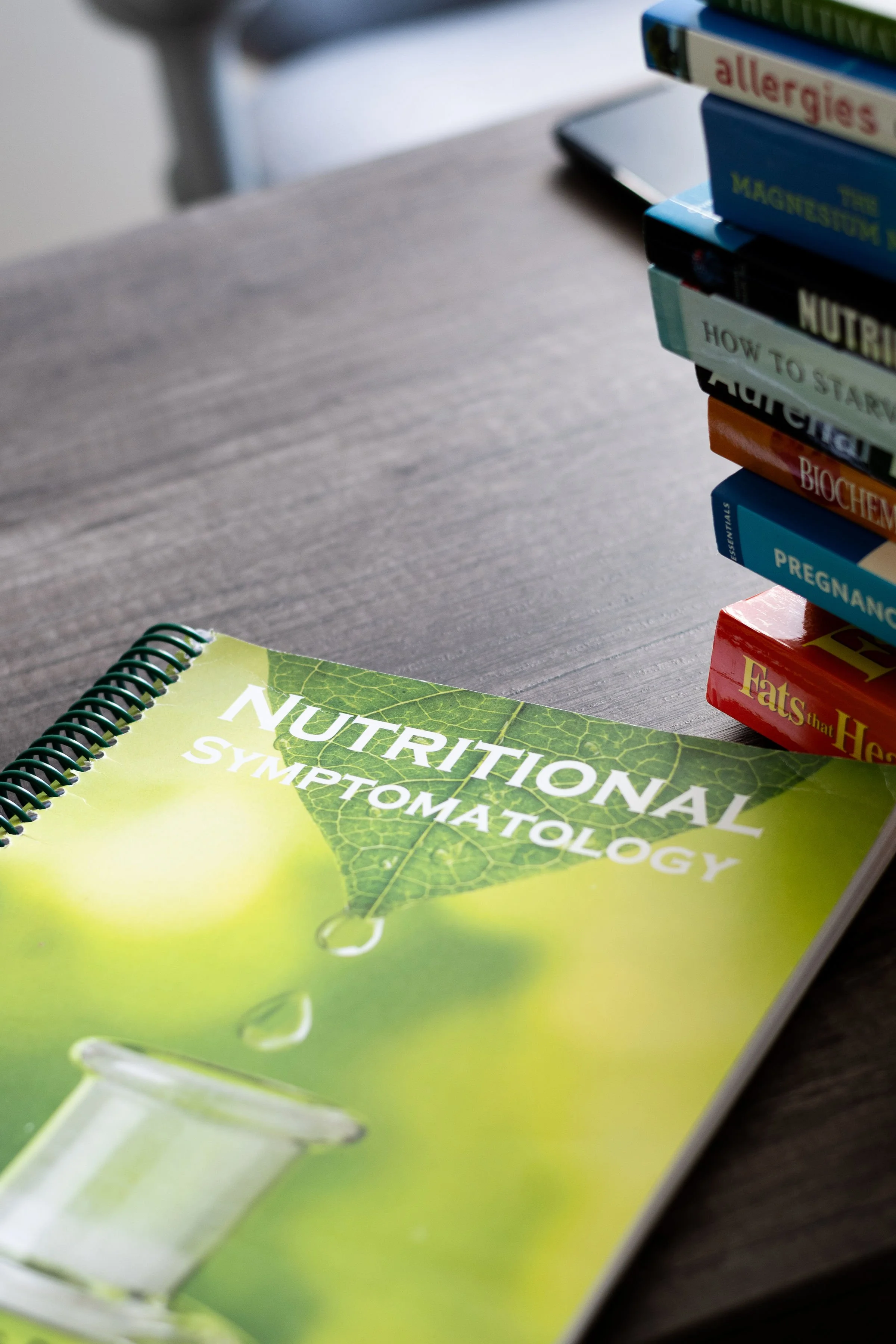 Close-up of a green nutrition book titled 'Nutritional Terminology' on a desk, with a stack of books about allergies, allergies, allergies, and pregnancy in the background.