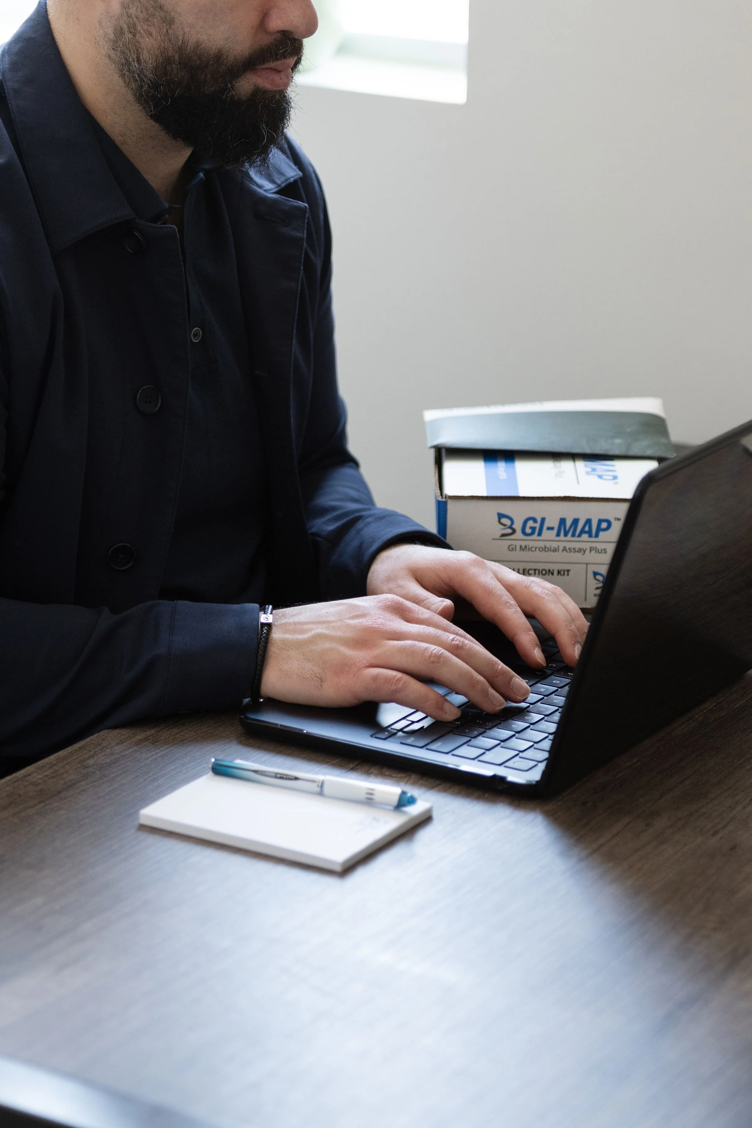 A person with a dark jacket and a beard, sitting at a wooden table, typing on a black laptop. A white notepad and a pen are on the table. Behind them, there is a box labeled "GI-MAP" with a dark green book on top. The background shows a white wall with a window letting in natural light.