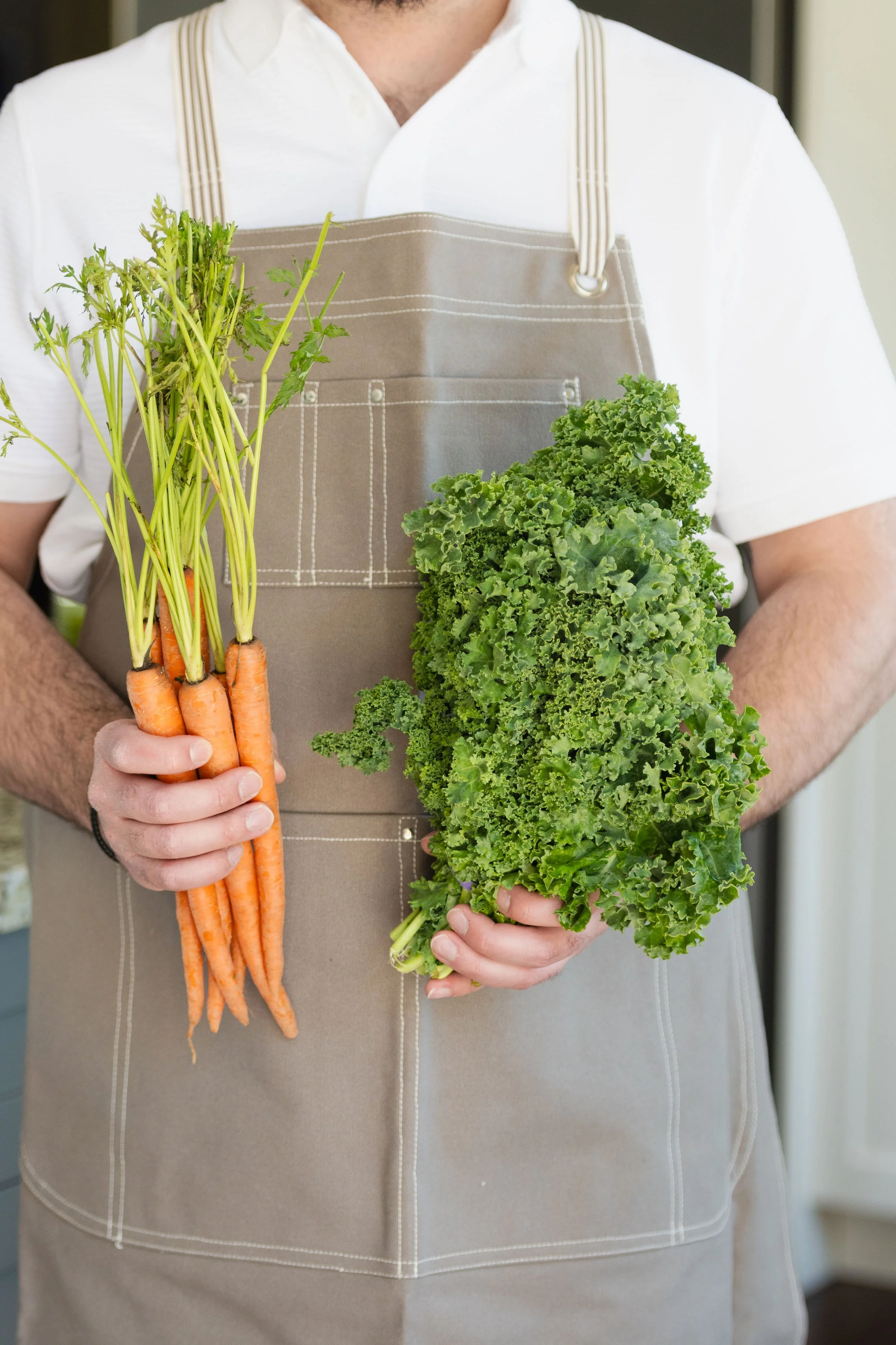 Person wearing a beige apron holding fresh carrots and curly kale.