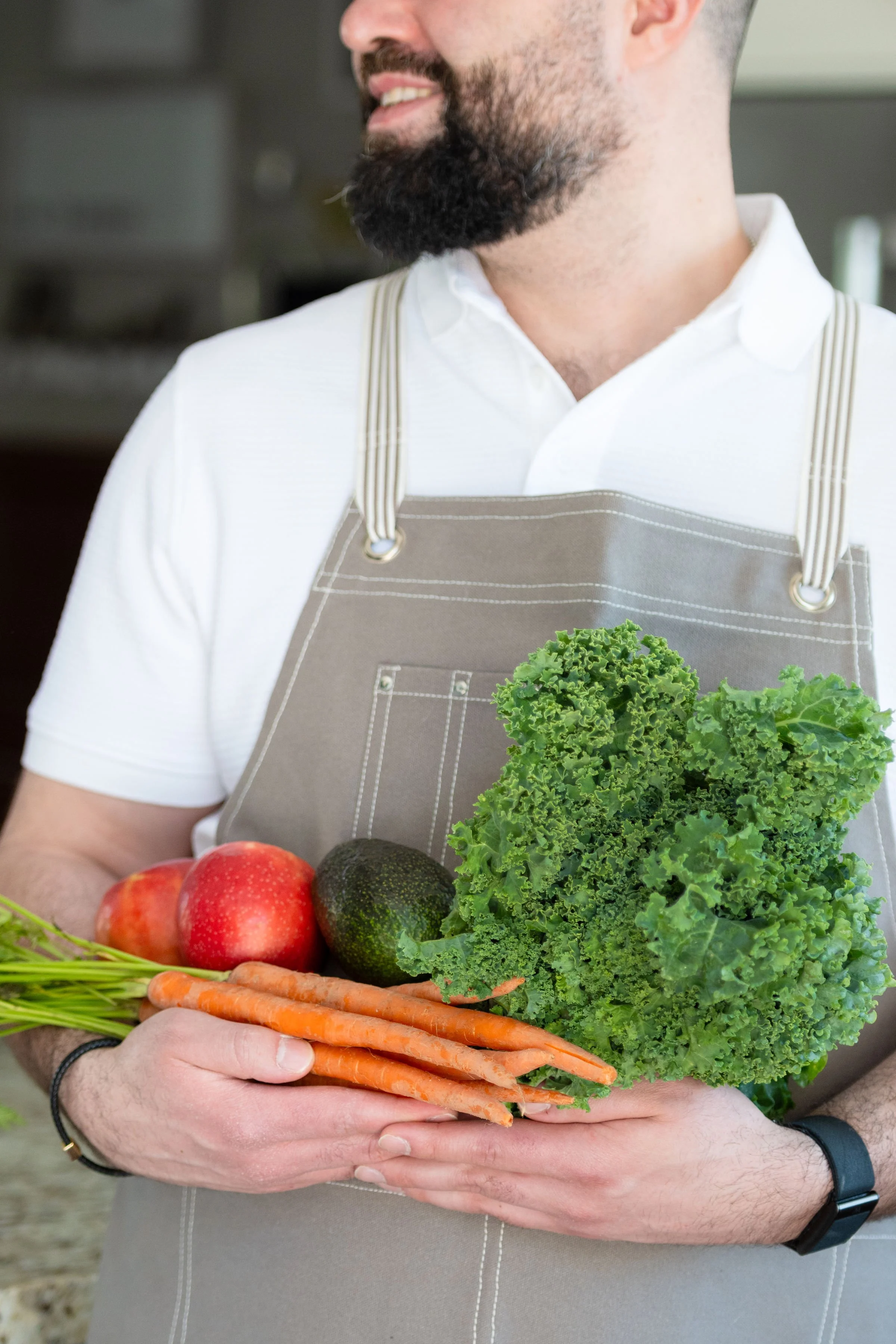 A man wearing a gray apron and white shirt holding a bunch of fresh vegetables including carrots, lettuce, apples, avocados, and carrots. The background is blurred.