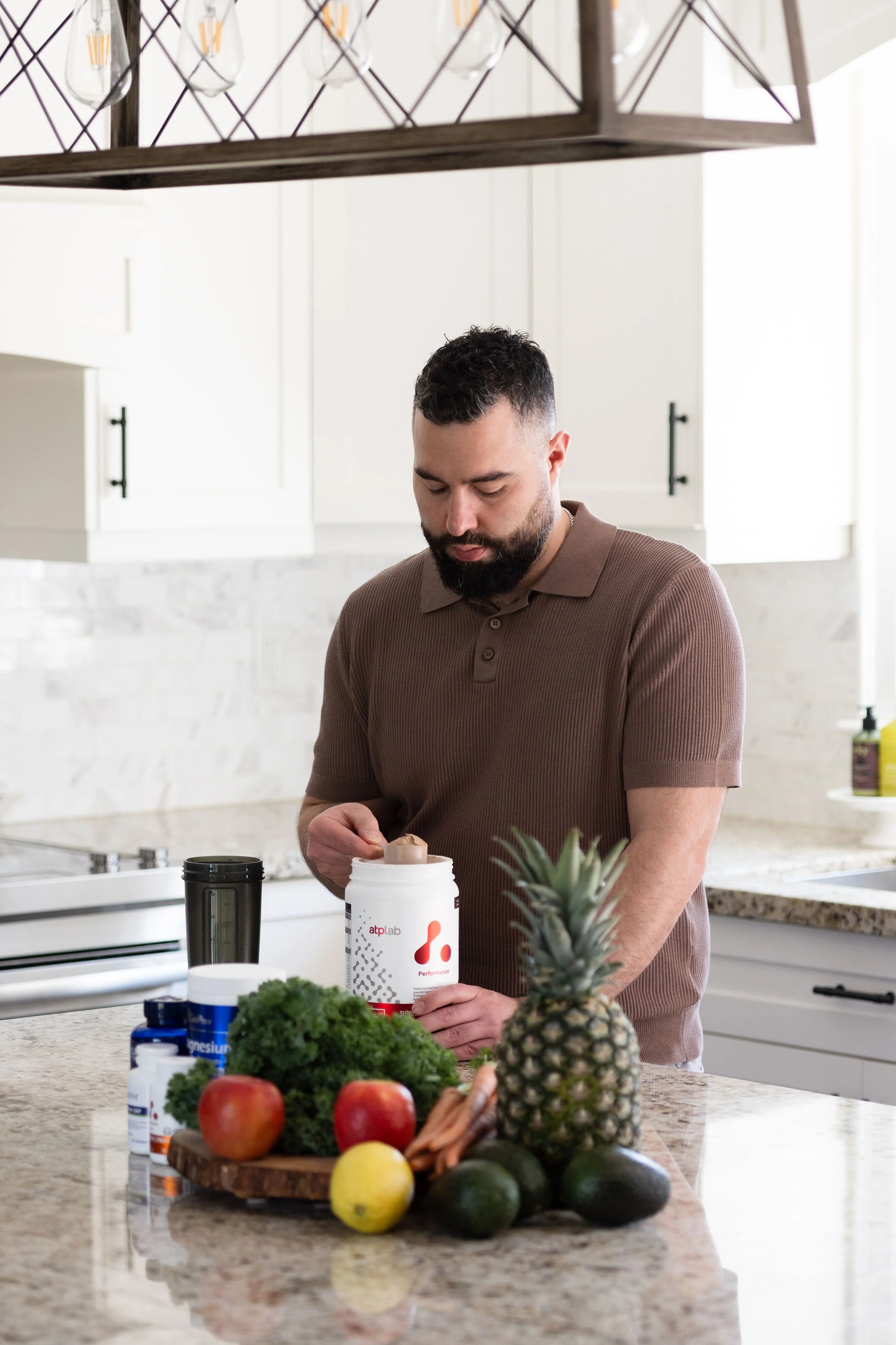 A man in a brown polo shirt opening a container of supplements in a modern kitchen. The kitchen has white cabinets, a granite countertop, and various fruits and vegetables including a pineapple, apples, avocados, kale, and lemons on the counter.