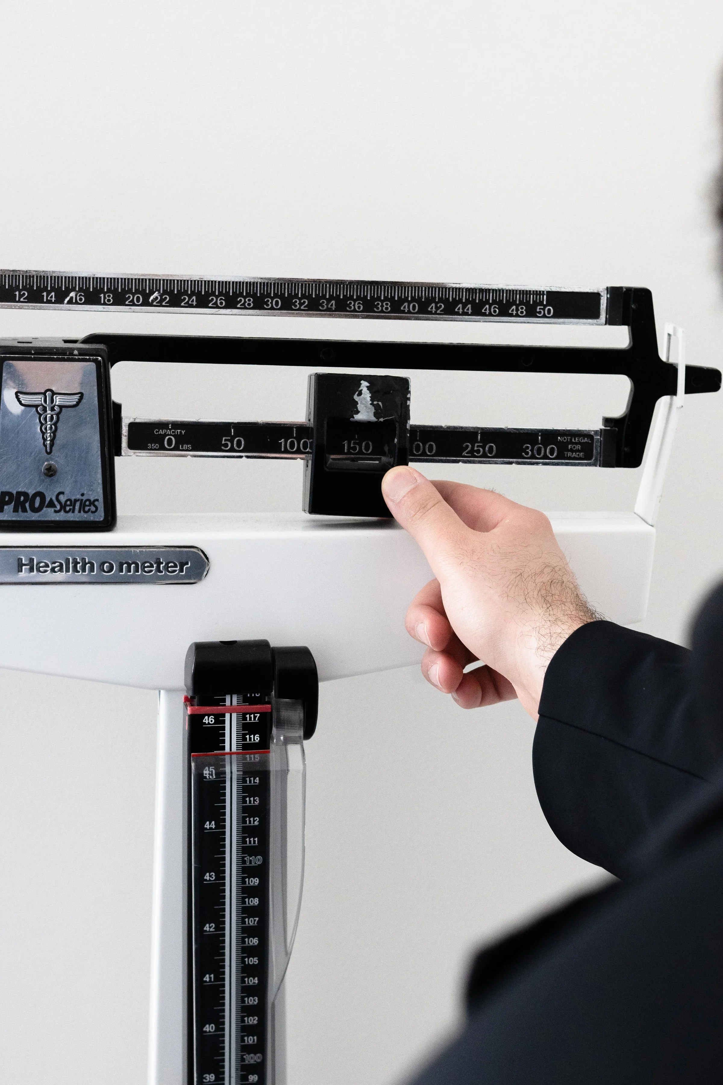 A person in a black suit is using a manual weighing scale with a black pointer, changing the weight measurement. The scale has a black and silver design with a capacity of 350 lbs and a height with a measuring tape. The background is plain white.