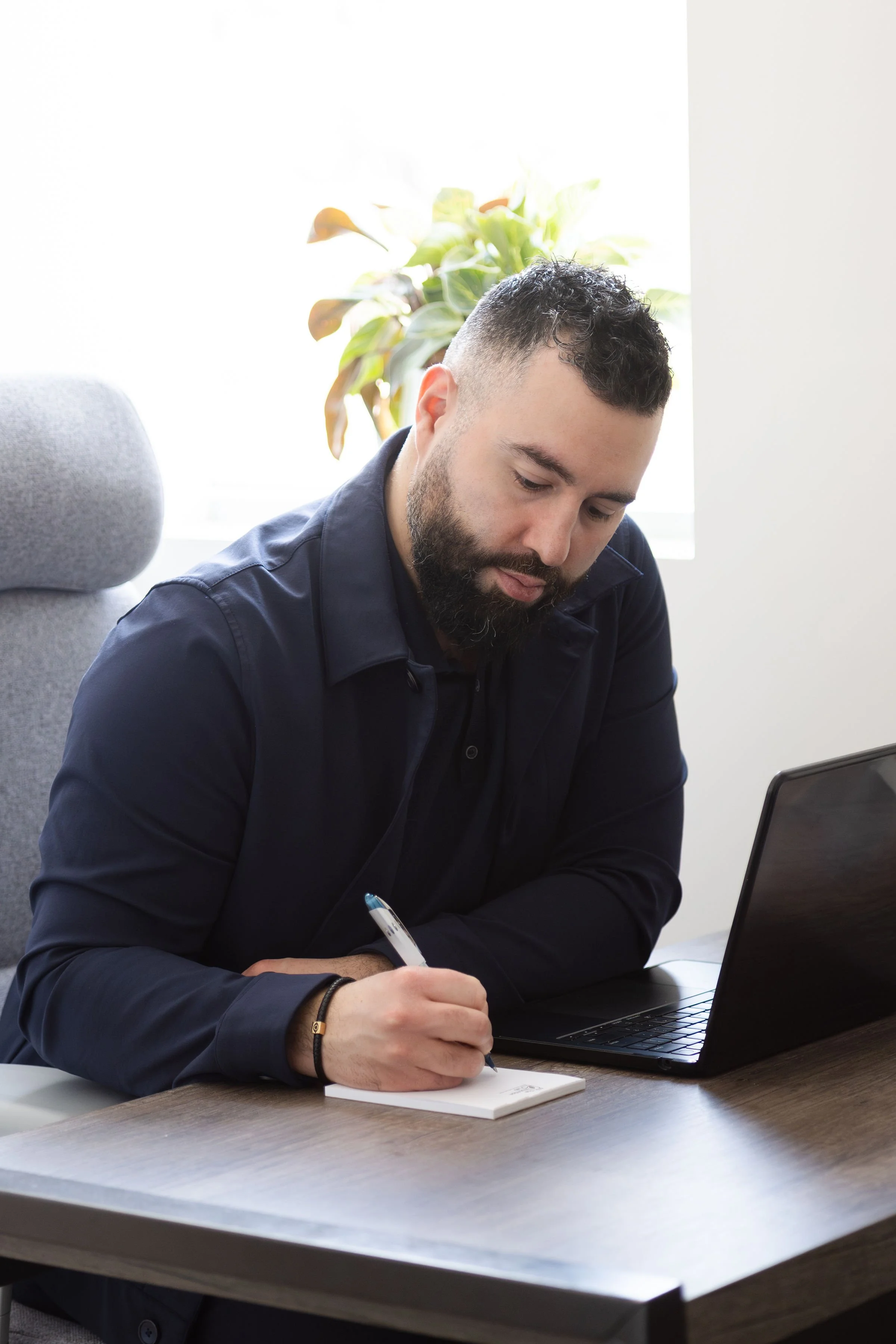 A man with dark curly hair and a beard sitting at a wooden table, writing in a notebook with a pen, with a laptop open in front of him and a plant in the background.
