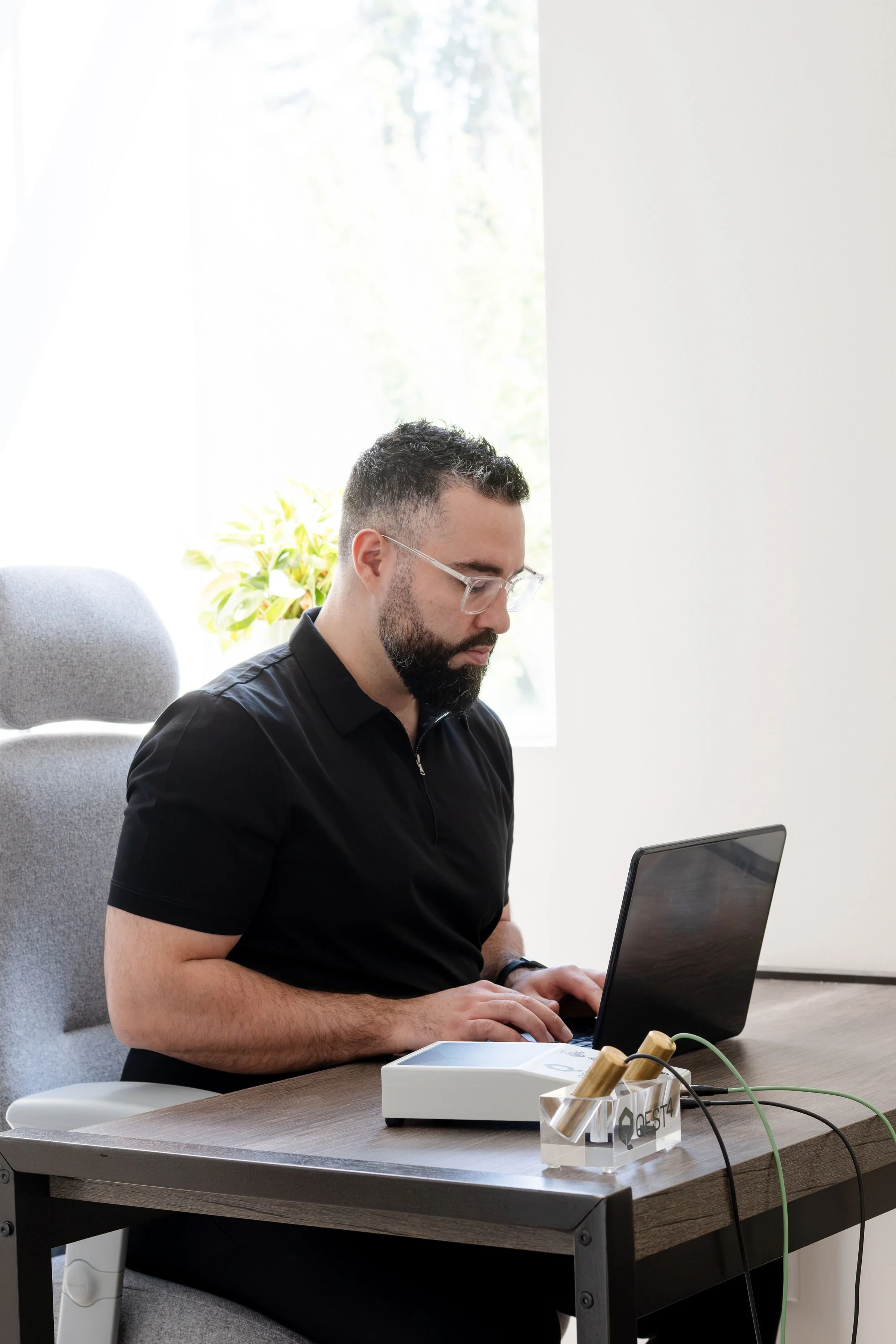 A man with short dark hair, beard, and glasses sitting at a desk working on a laptop. There is a window with natural light and a plant in the background.