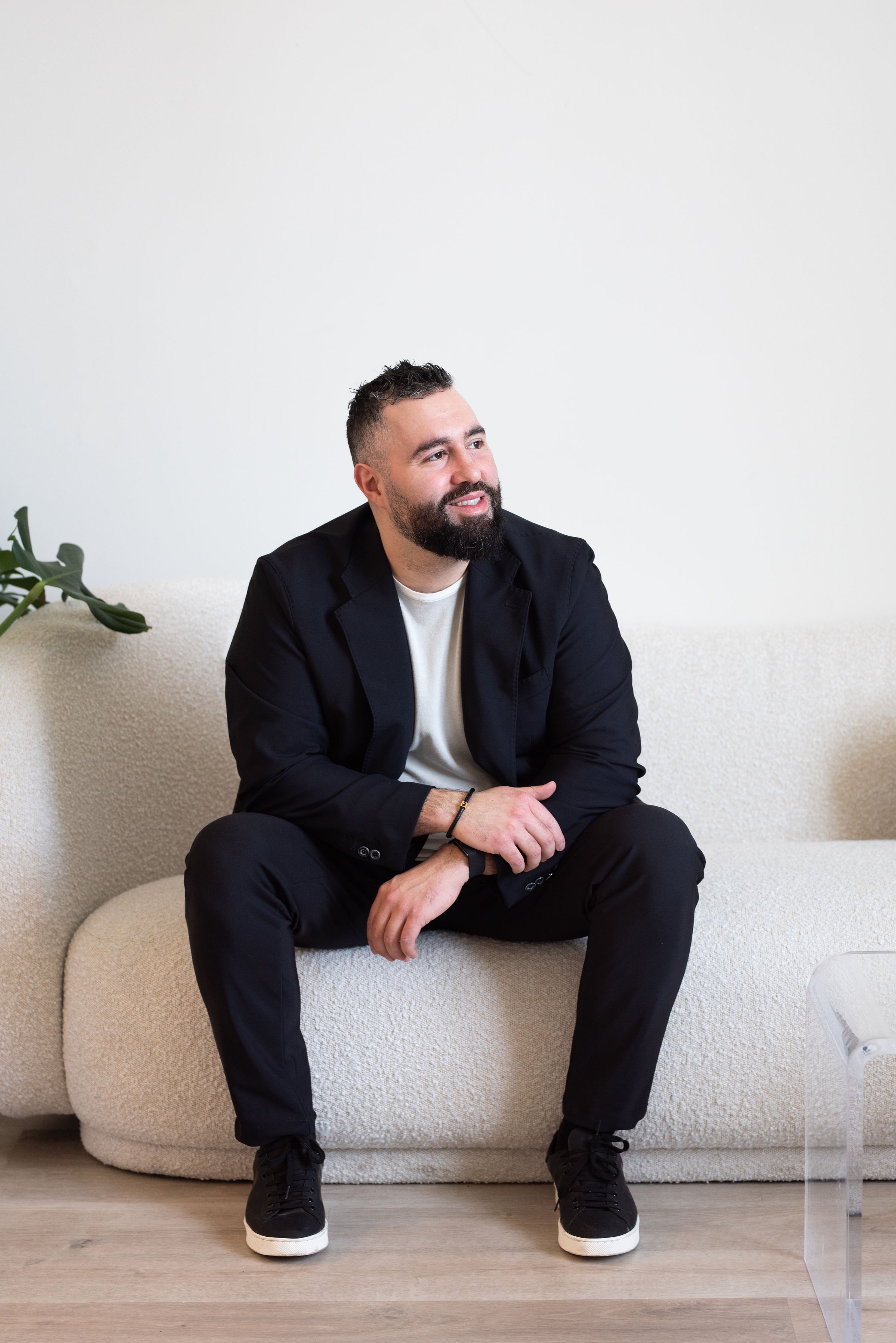 A man with a beard, wearing a black blazer, white t-shirt, black pants, and black sneakers with white soles, sitting on a light-colored textured sofa in a room with wooden flooring. A green plant is partially visible in the background.