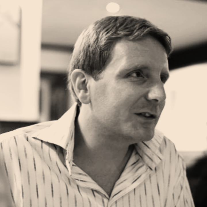A black and white photo of a man with short hair, wearing a striped collared shirt, looking slightly to the right and speaking.