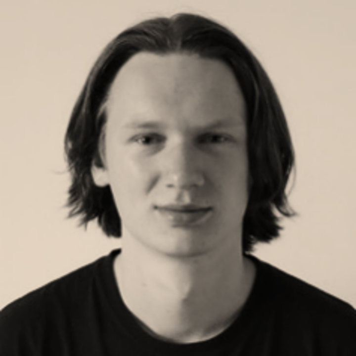 Black and white portrait of a young man with shoulder-length hair wearing a black shirt, against a plain background.