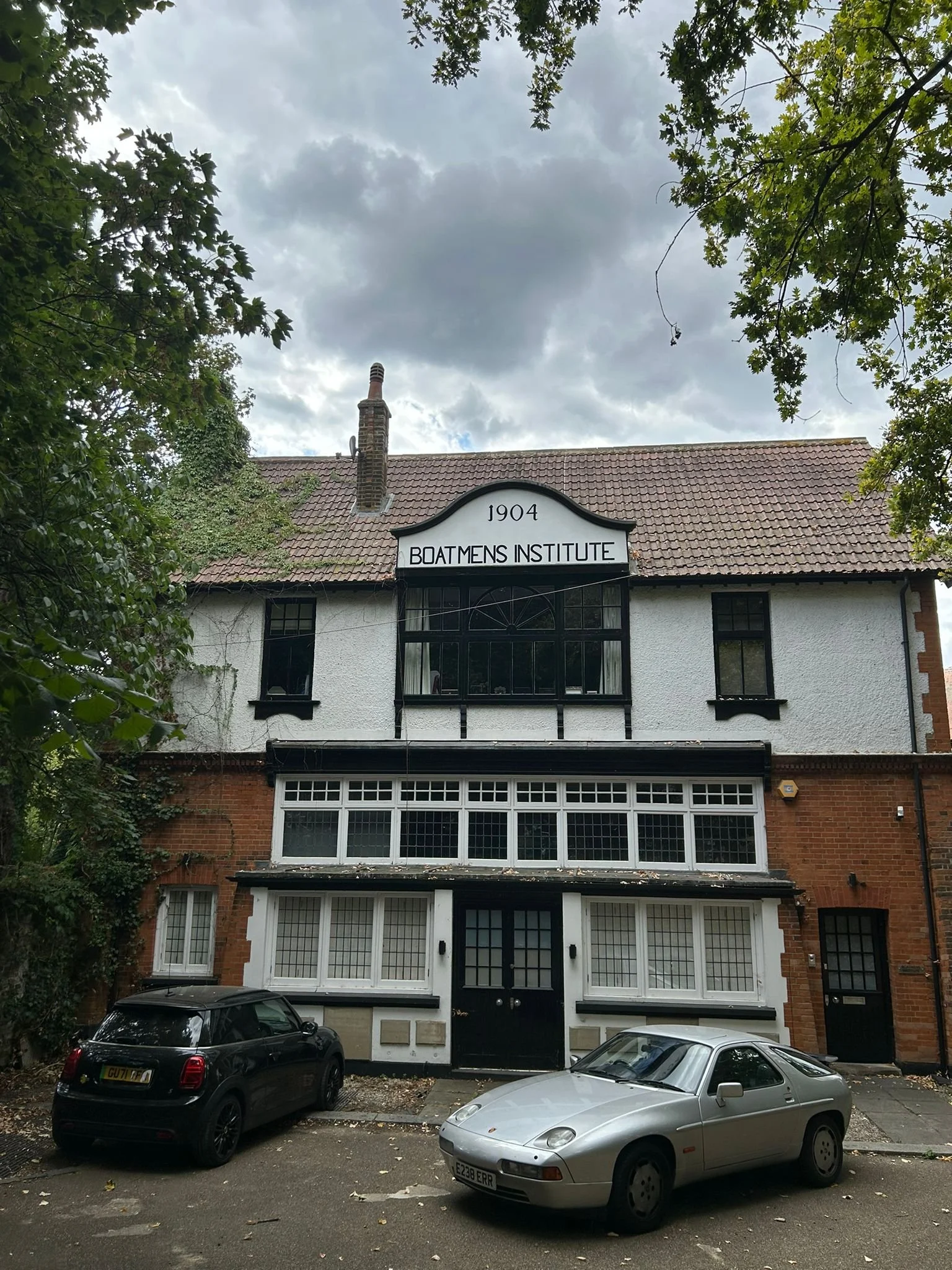 Building with a sign that reads 'Boatmen's Institute,' a brick chimney, trees framing the top, and three cars parked in front. Brentford, The Butts.