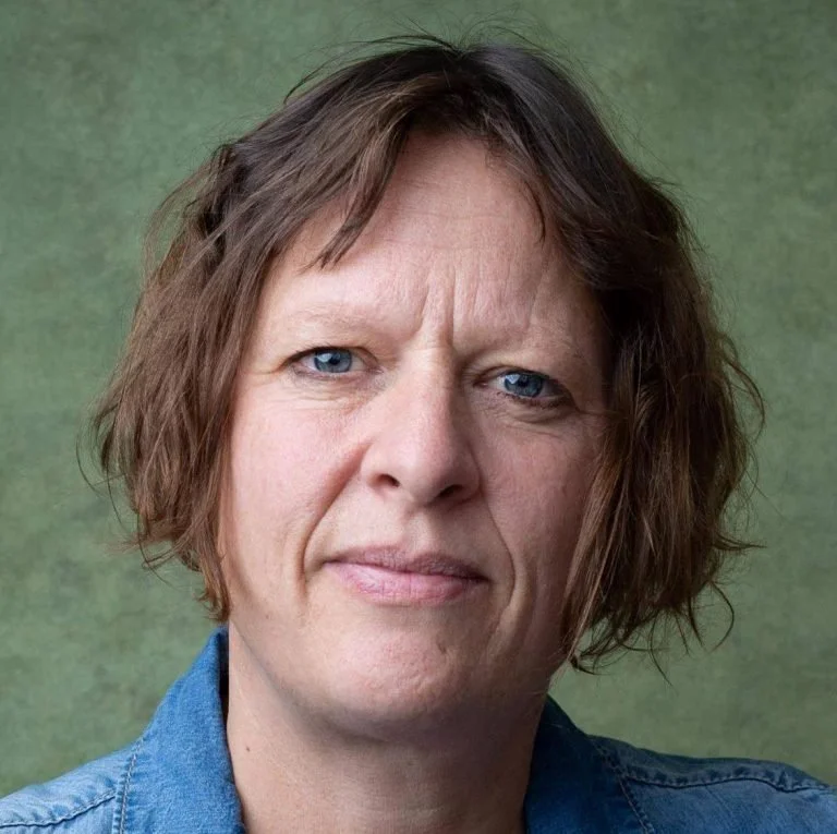 Close-up portrait of a woman with short, wavy brown hair and blue eyes, wearing a denim shirt, against a green background.