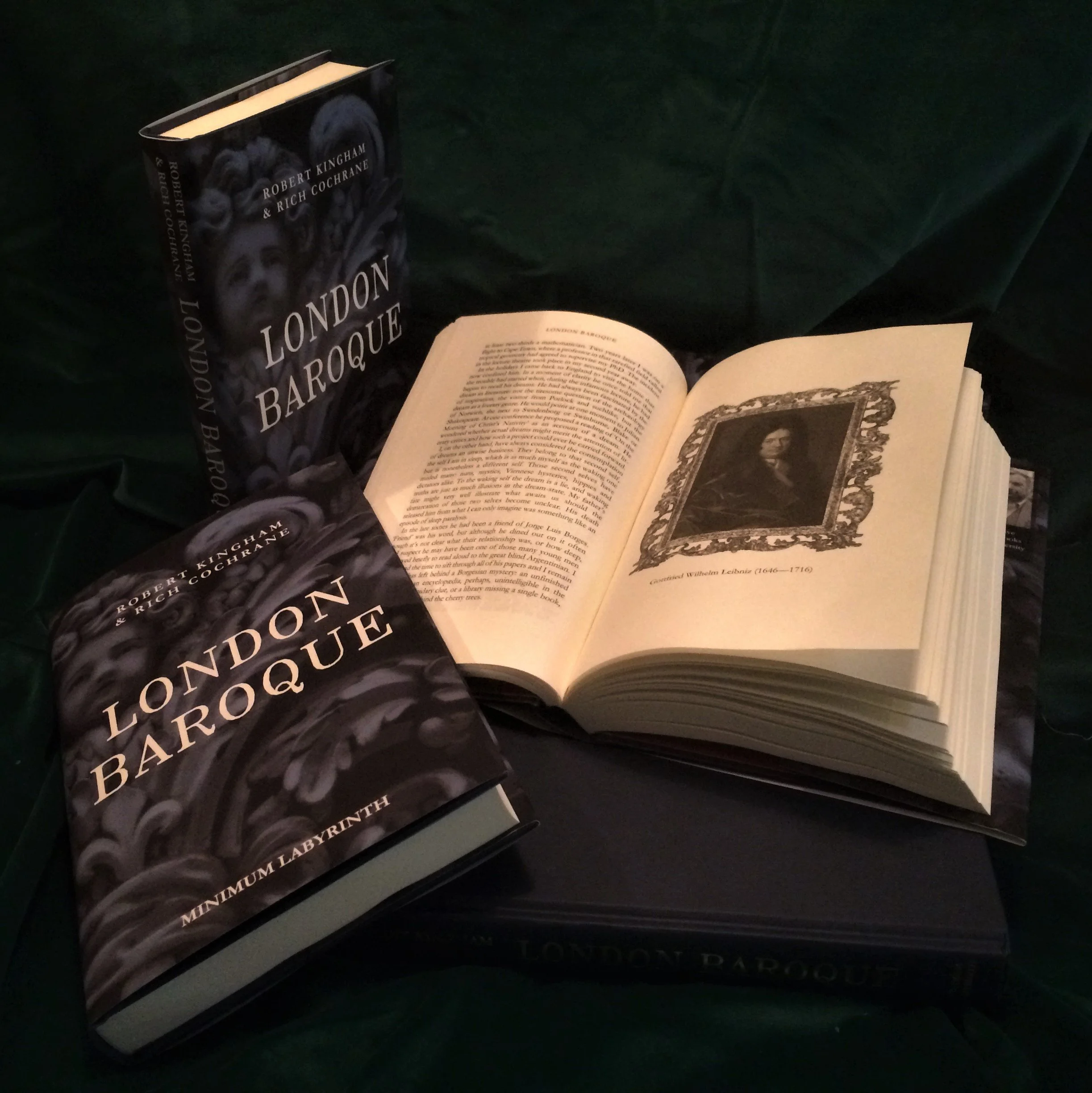 Photo of two books, one closed and one open, on a dark surface. The closed book is titled 'London Barroque' with a dark cover featuring a portrait. The open book displays a black-and-white illustration of a woman framed in an ornate border, along with text.
