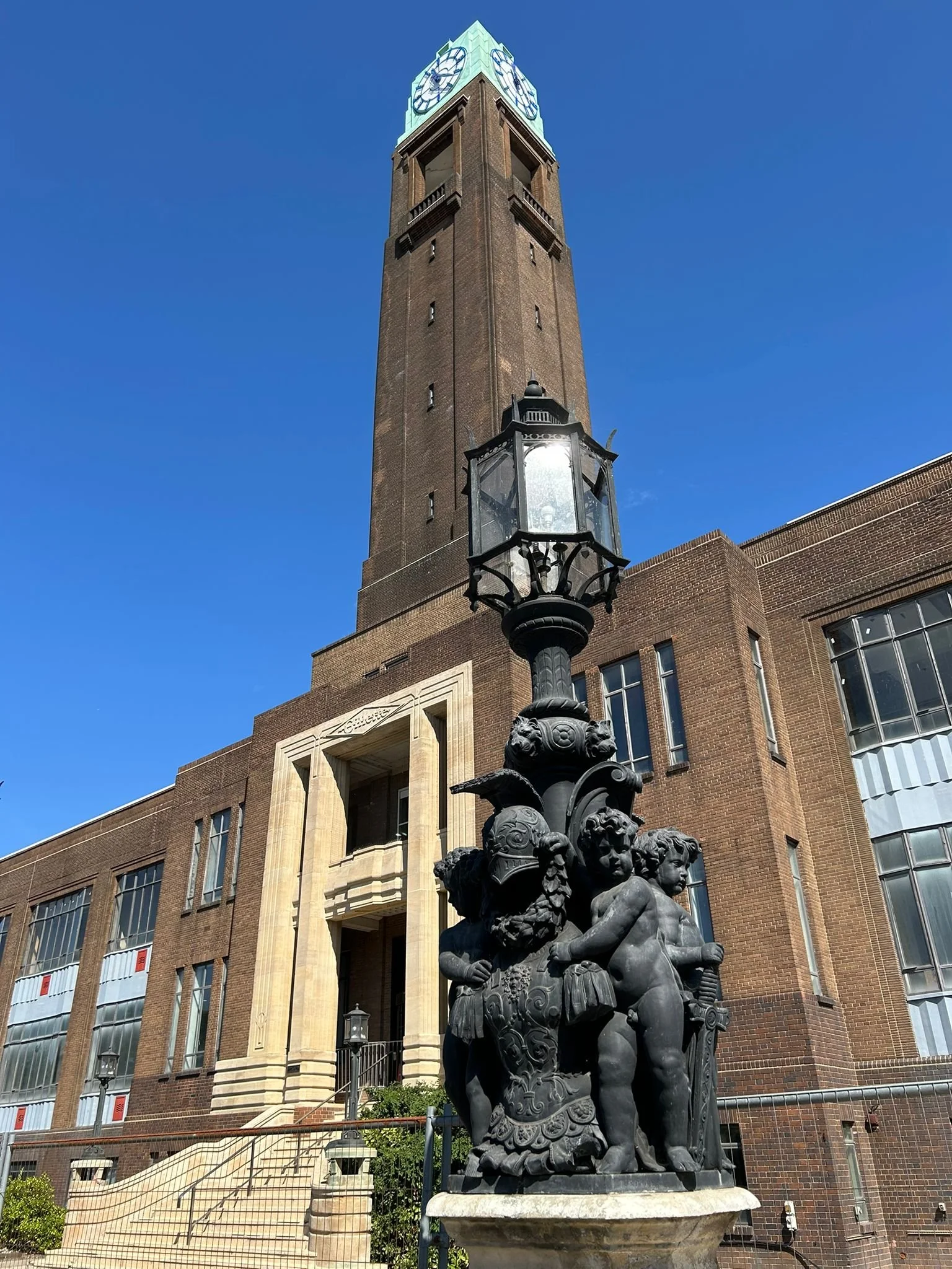 Statue of cherubic children around a shield, with a large clock tower in the background, against a clear blue sky. The Gillette Building in Brentford.