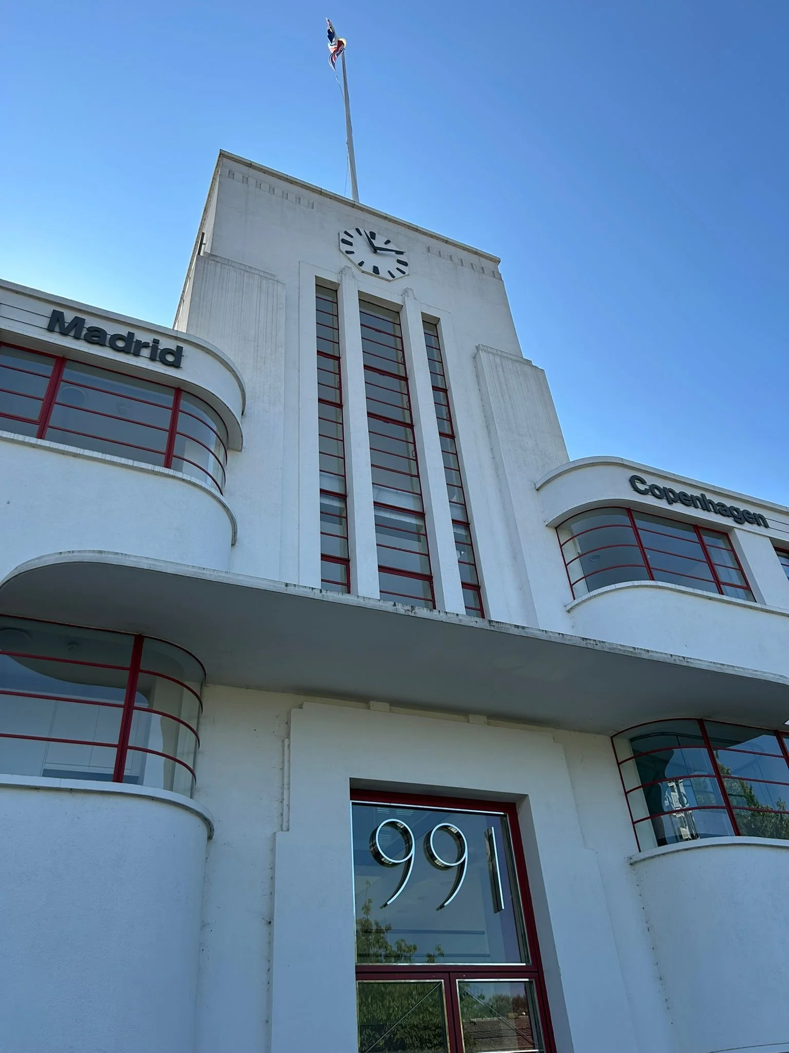A white building with curved balconies and red window frames, featuring a clock tower with a Union Jack flag on top. The sign reads 'Madrid' and 'Copenhagen'. The Golden Mile, Brentford. JCDecaux building, Currys headquarters from 1936.