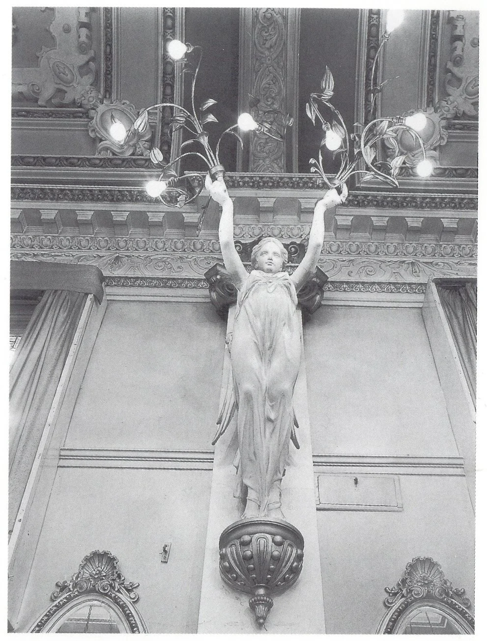 A black and white photo featuring a statue of a woman with arms raised, holding a decorative chandelier with lights. A ‘Clerkenwell angel’ inside Finsbury Town Hall.