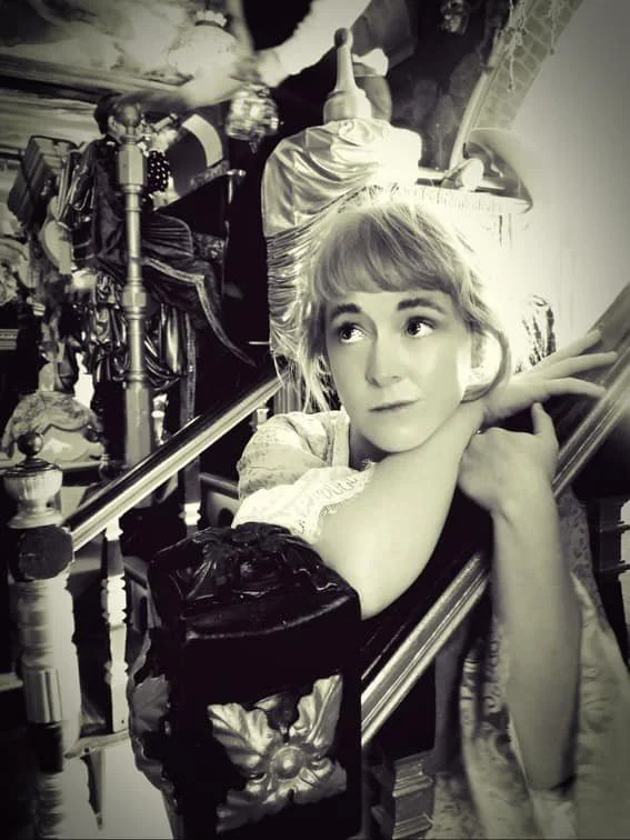 Alice Merivale as Nell Gwyn, resting her chin on her arms on a staircase, looking thoughtfully to the side, in Sarastro restaurant, filled with vintage furniture and decor.