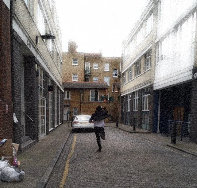 The doomed young man in spectacles running down a narrow urban street in Clerkenwell, surrounded by parked cars and multi-storey buildings.