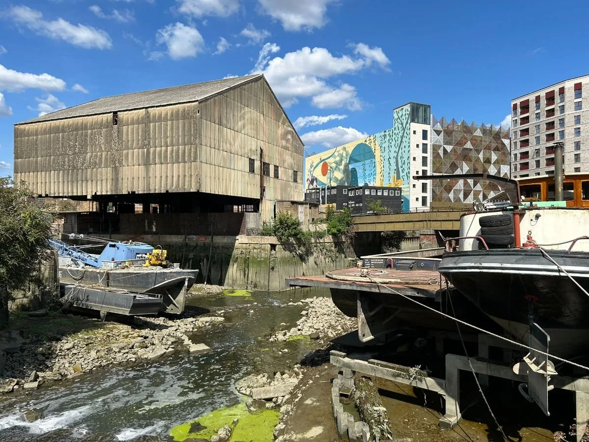 Brentford. A daytime scene of boats docked alongside an industrial building and colourful modern buildings in an urban canal area.