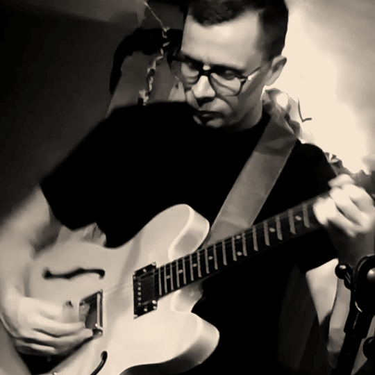 A man wearing glasses playing an electric guitar in a recording studio.