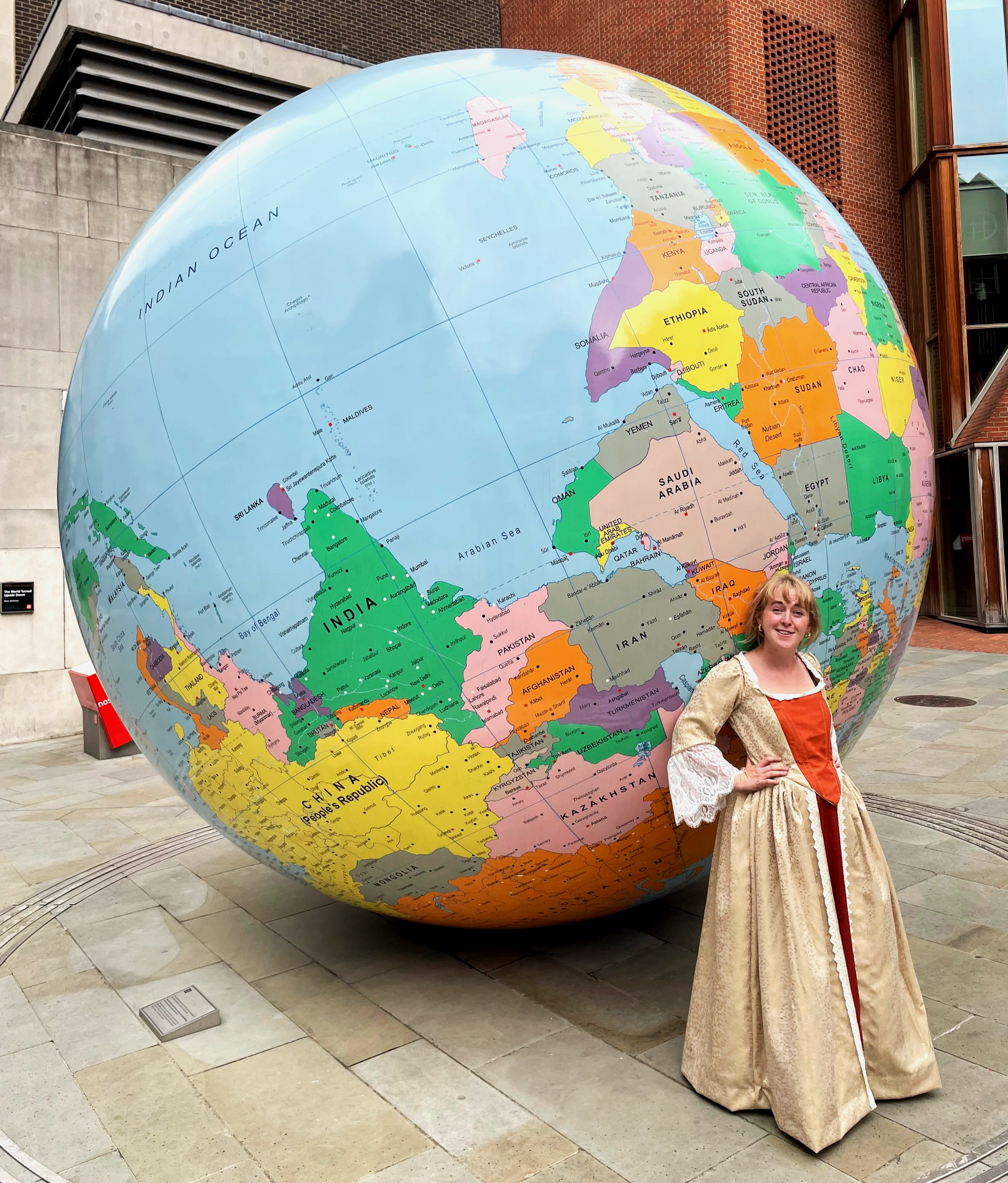 Alice Merivale as Nell Gwyn. A woman dressed in a historical costume standing next to a large globe outdoors near a building with brick and glass walls.