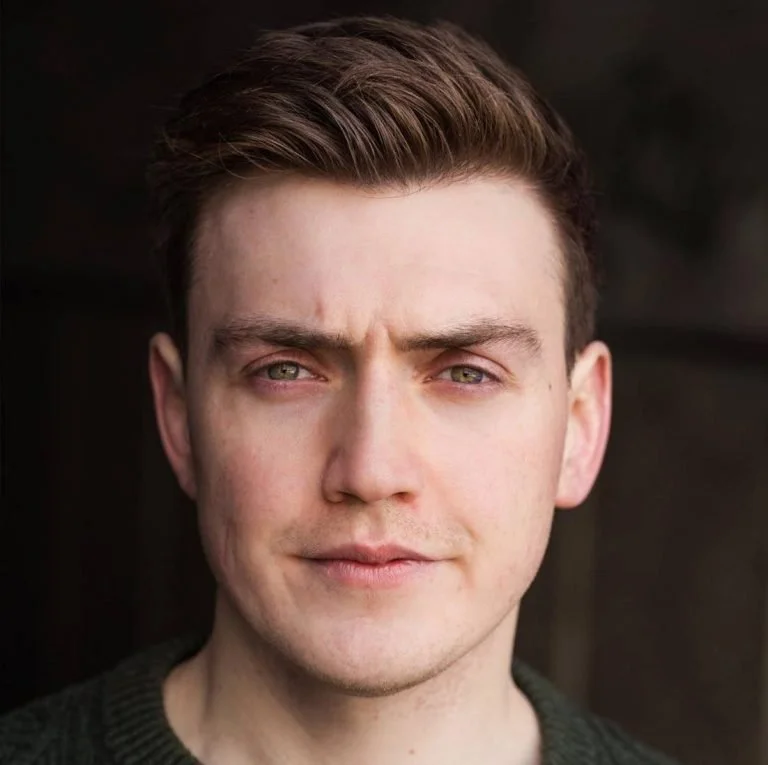 Close-up of a young man with short brown hair and light green eyes, wearing a dark sweater, looking directly at the camera with a neutral expression.