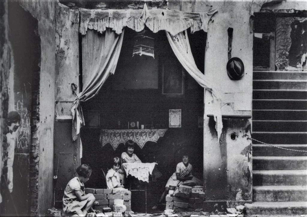 The ‘little housewives of Sadler’s Wells’, 1923. A black and white photo showing a small group of children sitting outside a damaged building, with torn curtains hanging, a staircase on the right, and old, worn walls.