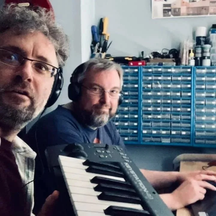 Two men wearing headphones sitting at a desk with a keyboard, in a room with a blue tool organizer and various supplies on the wall and shelves.