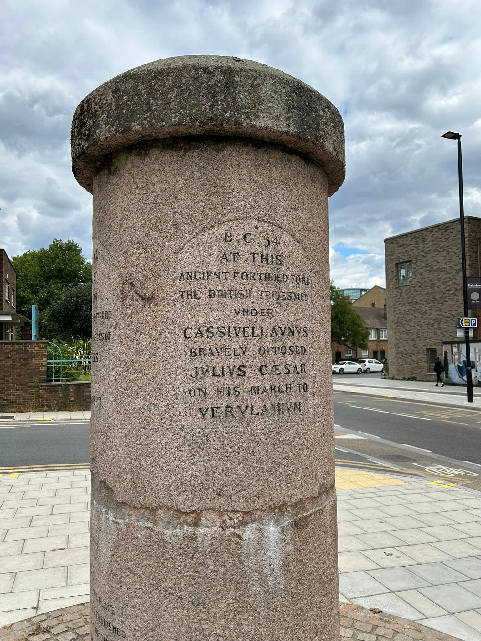 A stone monument with an inscription, located in an urban area with buildings, trees, a parking lot, and pedestrians nearby, under a cloudy sky. The Brentford Monument.