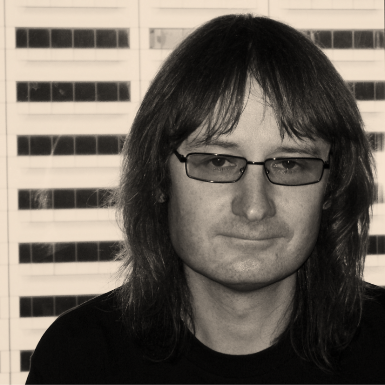 A person with long hair, glasses, and a slight smile standing in front of a building with multiple windows.