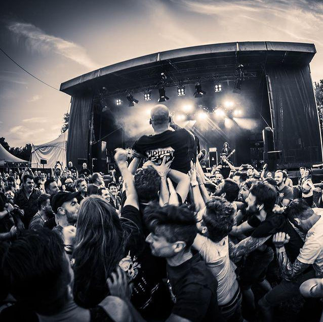 Band performing on stage at an outdoor concert with a crowd in front, many raising their hands and a person crowd surfing.