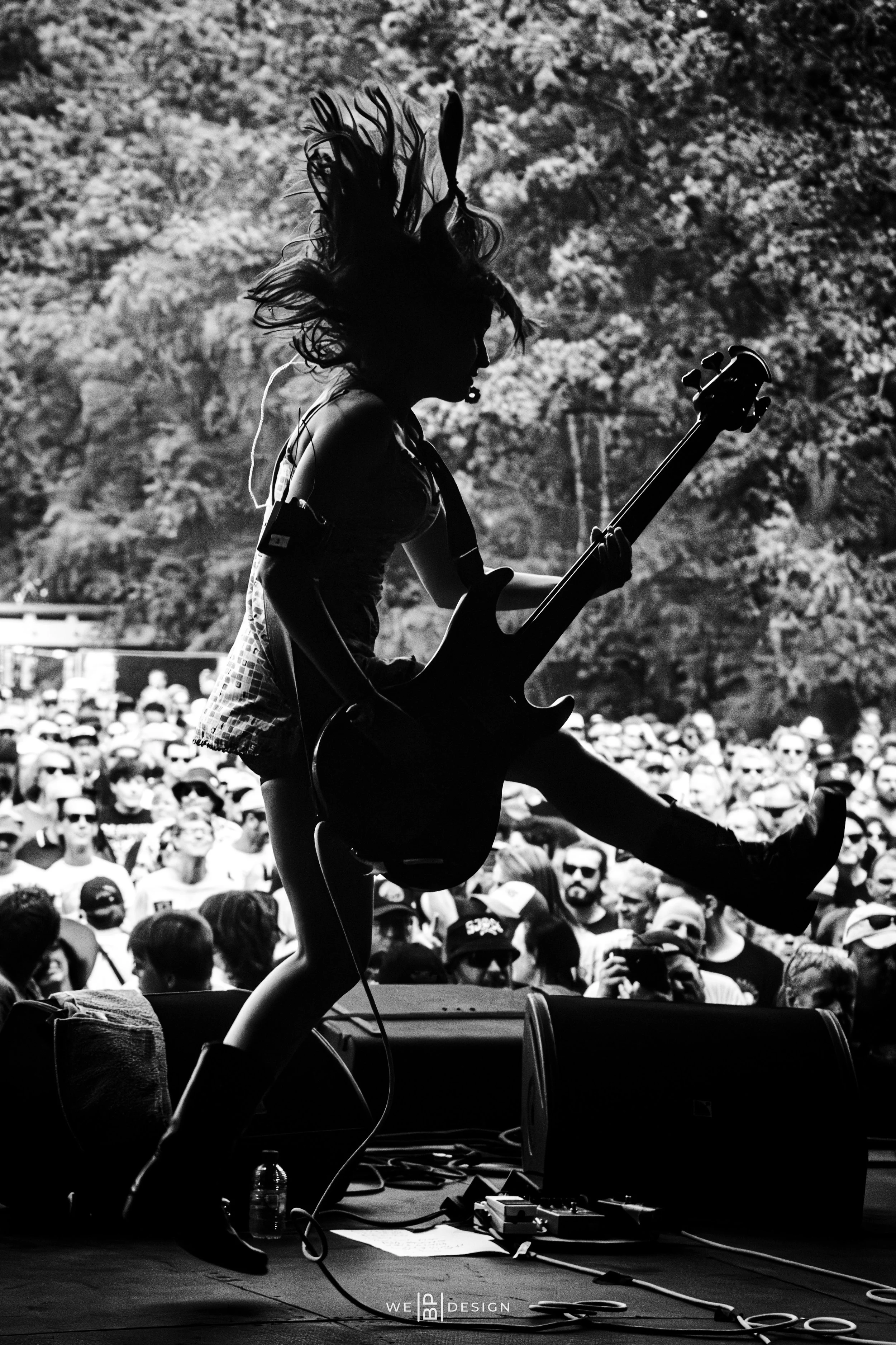 A silhouette of a woman playing an electric guitar on a stage at an outdoor concert, with a large crowd and trees in the background.