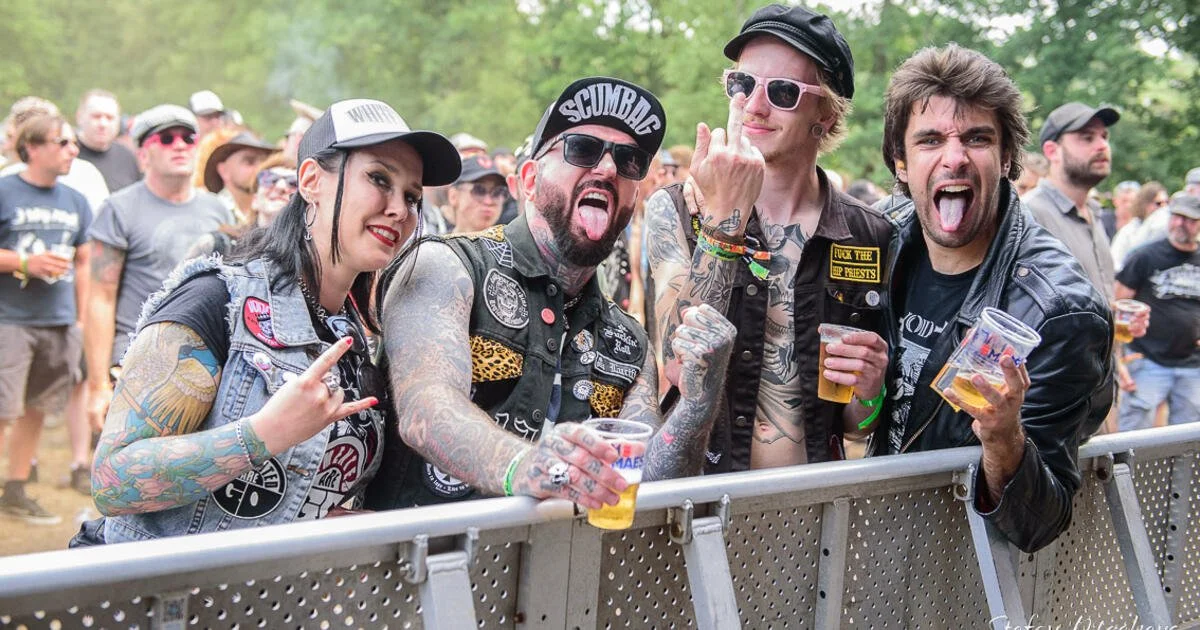 Four friends at a music festival, leaning on a barrier, holding drinks, making rock hand signs, and sticking out their tongues.