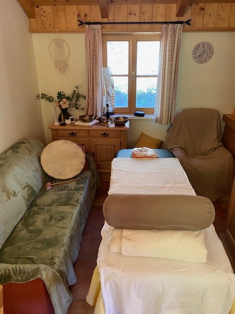 Gemütliches Wohnzimmer mit einem Sofa, einem Sessel, einem Tisch, einem Fenster, Vorhängen, Uhren, und Dekorationsgegenständen auf einem Sideboard.