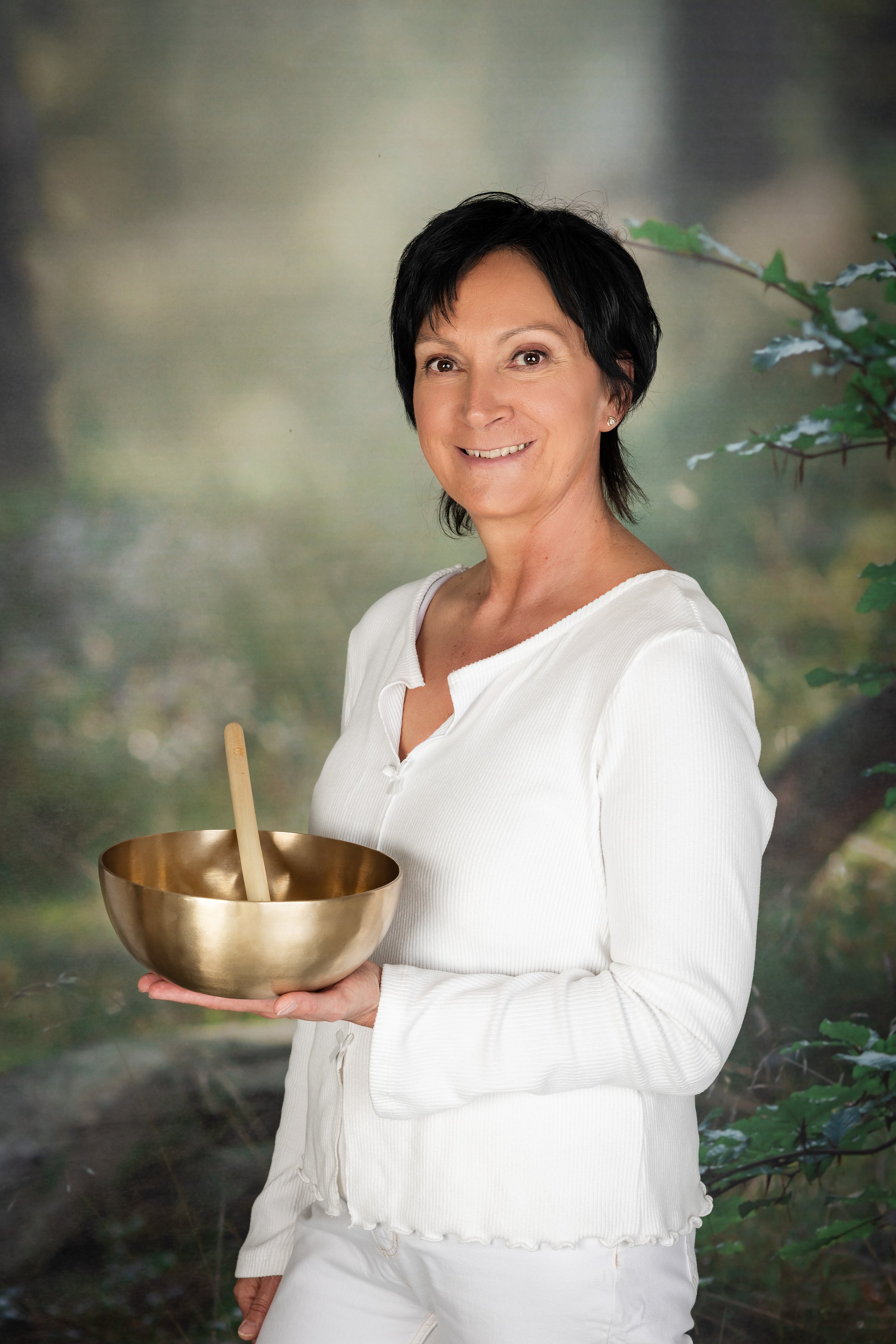 Eine Frau mit kurzen schwarzen Haaren trägt ein weißes Oberteil und hält eine goldene Klangschale mit einem hölzernen Schlägel. Im Hintergrund befindet sich eine bewaldete Szene.