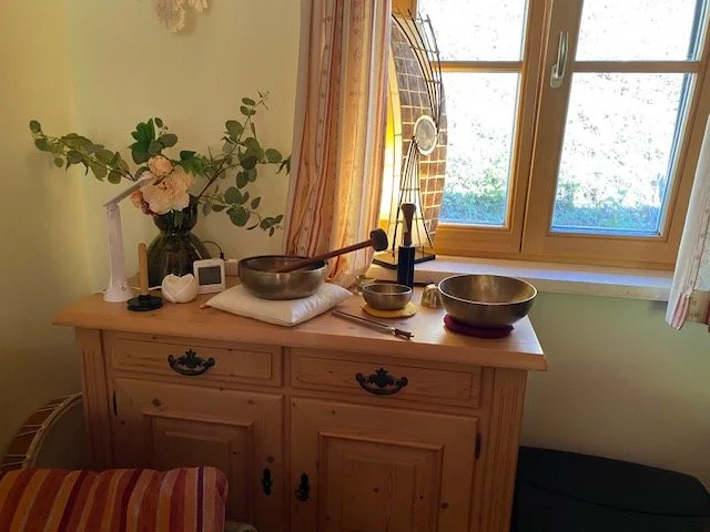 Ein Holzsideboard mit verschiedenen Gegenständen, darunter eine Vase mit pinken und weißen Blumen, ein Schneidebrett, eine weiße Kerze, eine Uhr, eine kleine Lampe, zwei runde Metallschalen und ein Chakra-Klangschale, neben einem Fenster mit Vorhänge