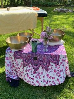 Ein Tisch im Garten mit drei Klangschalen, einem Blumenvase mit Blumen, einer grünen Flasche, gedeckt mit einer lila und weißen Tischdecke.
