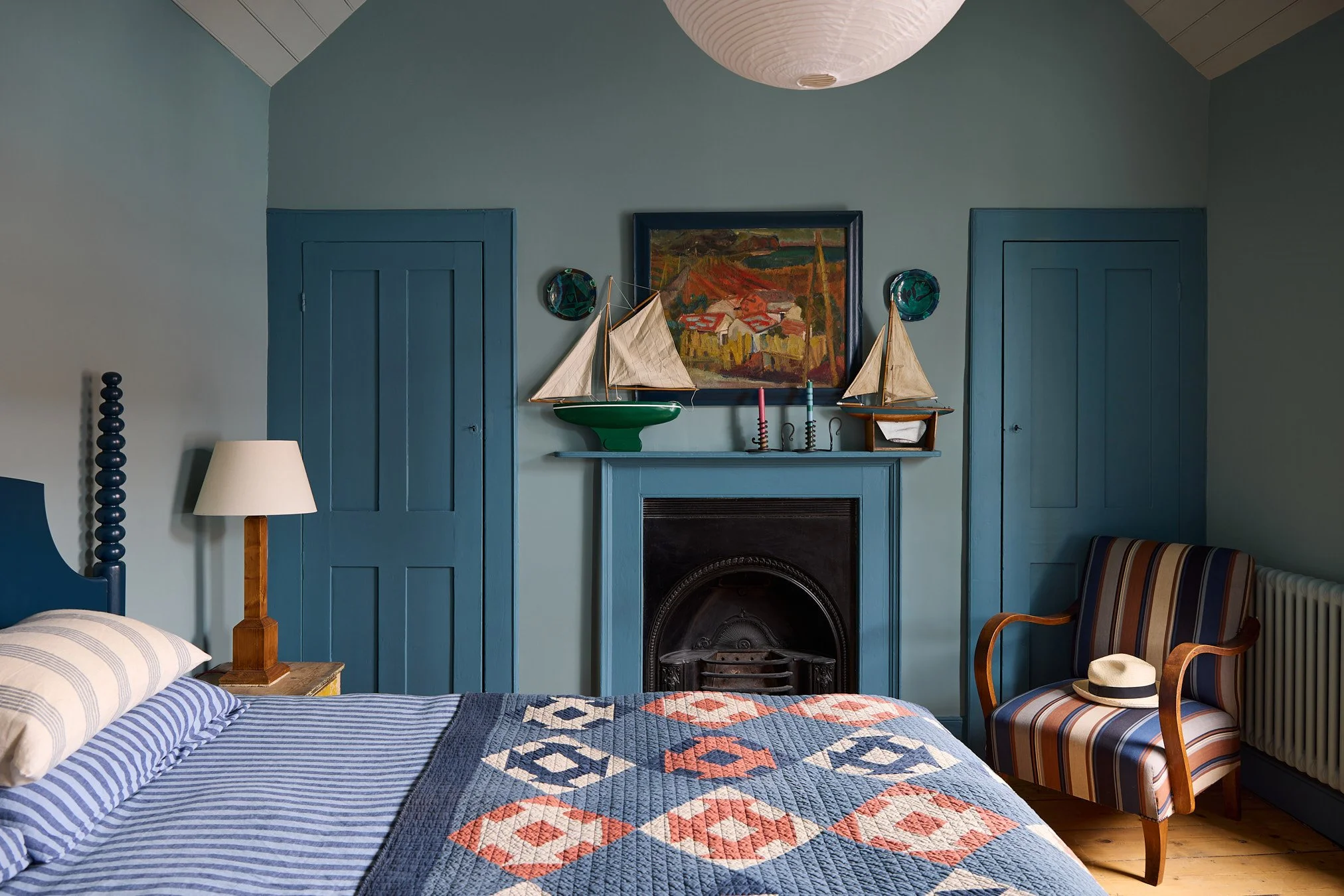 A cozy Georgian  Cottage bedroom with a blue color scheme, featuring a bed with striped bedding, a wooden nightstand, a lamp, and a fireplace with nautical-themed decorations and artwork on the wall.