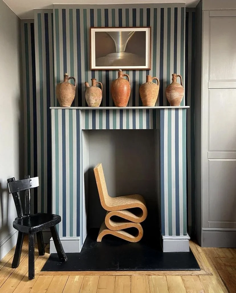 A decorative fireplace with five ceramic vases on the mantel, striped wallpaper, a framed picture above, a modern wooden chair with a looped design inside the fireplace opening, and an additional small black chair to the side, all on a wooden floor.