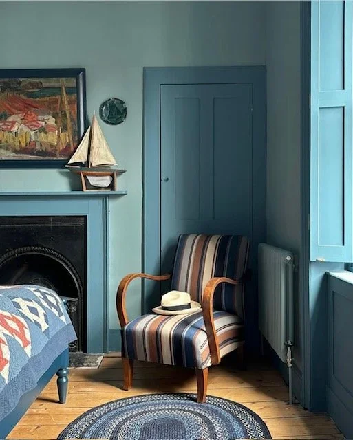 A cozy bedroom corner with a striped armchair, a hat on it, a blue painted wall, a fireplace, a framed painting above the fireplace, a wooden model sailboat on the mantle, a small round clock on the wall, a blue door, window shutters, a radiator, and a braided rug on wooden floor.