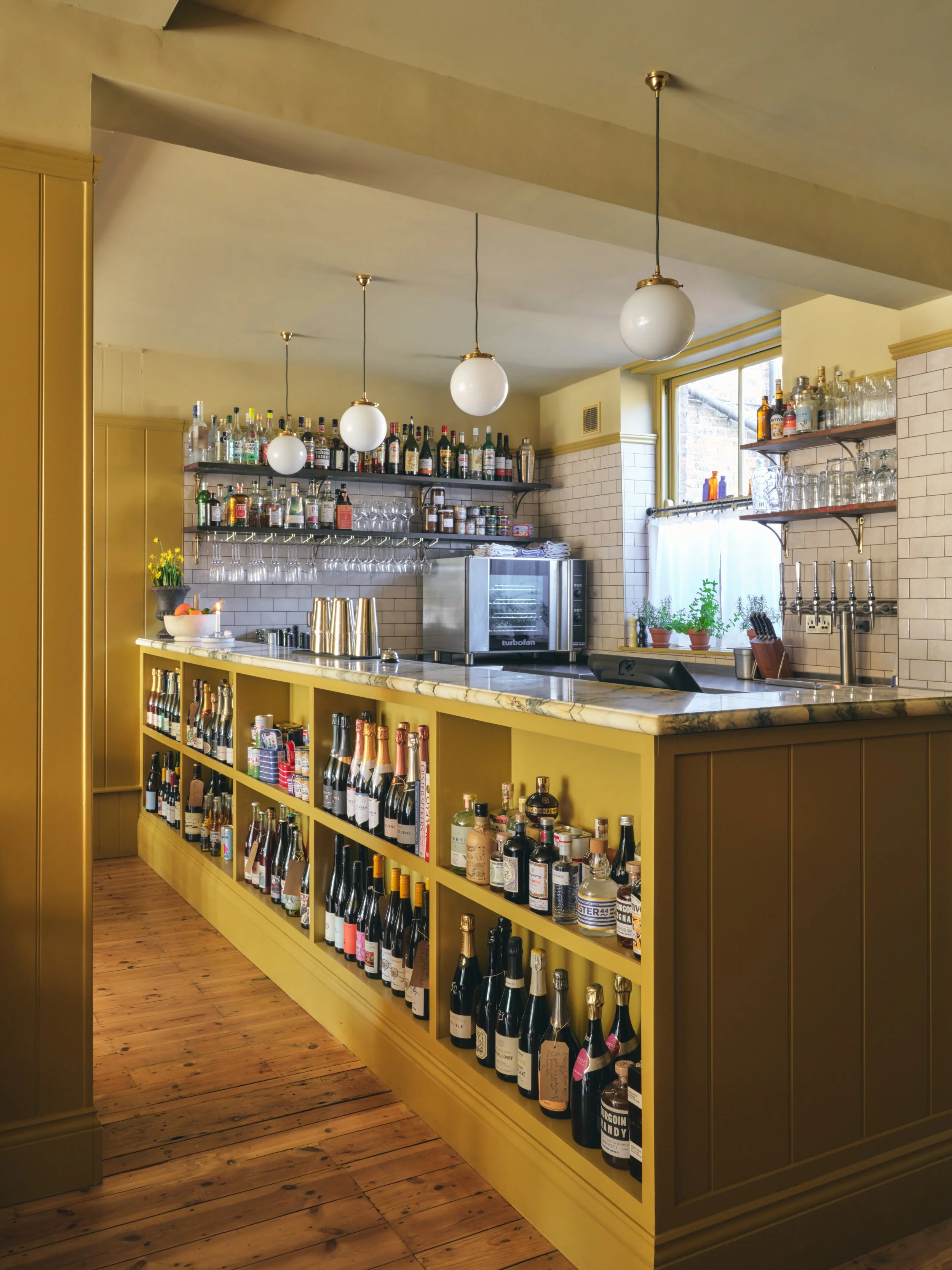 A bar with shelves of various bottles of alcohol, wine, and glasses, yellow cabinetry, a marble countertop, and a window with plants and glassware.