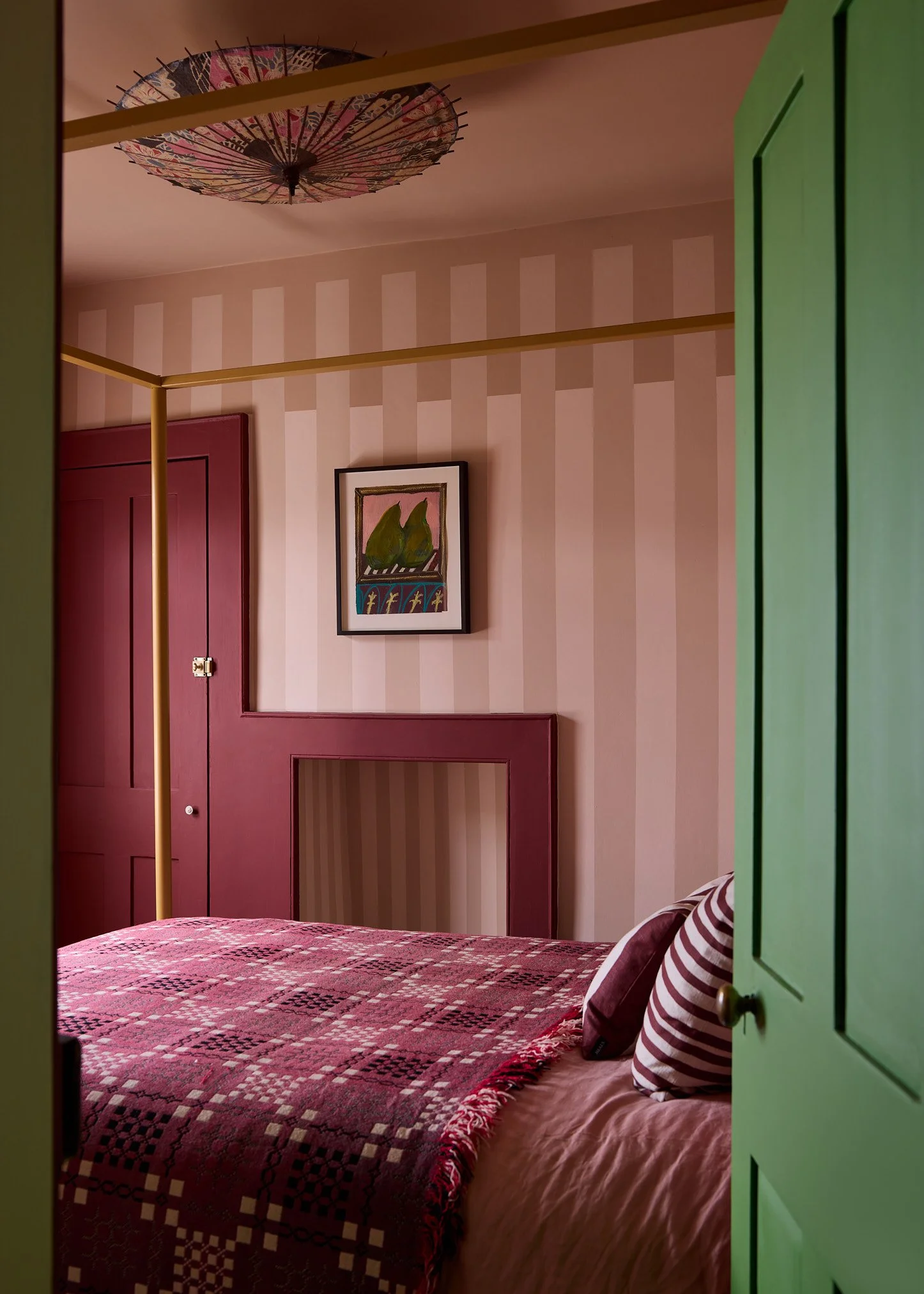 View of a bedroom through a partially open green door, showing a bed with a patterned red, black, and white bedspread, a pillow with red and white stripes, a framed artwork of green pears on a pink background on the wall, and a pink and cream striped wallpaper.