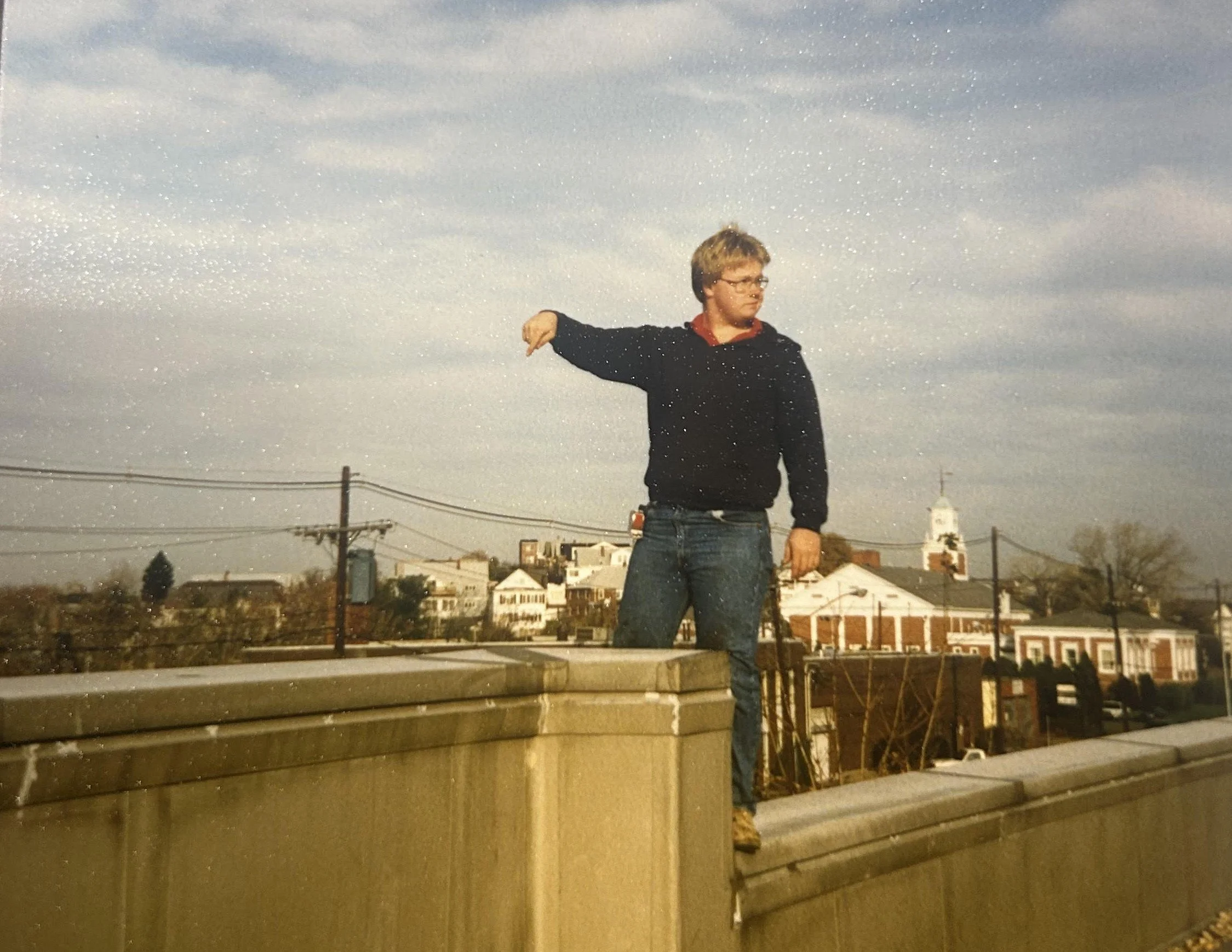 A young man with glasses, wearing a black hoodie and jeans, standing outdoors on a rooftop or balcony during dusk or early evening. He is facing to the right with his left arm extended outward and his index finger pointing downward. The background shows a cloudy sky, power lines, and a neighborhood with houses and church steeples.