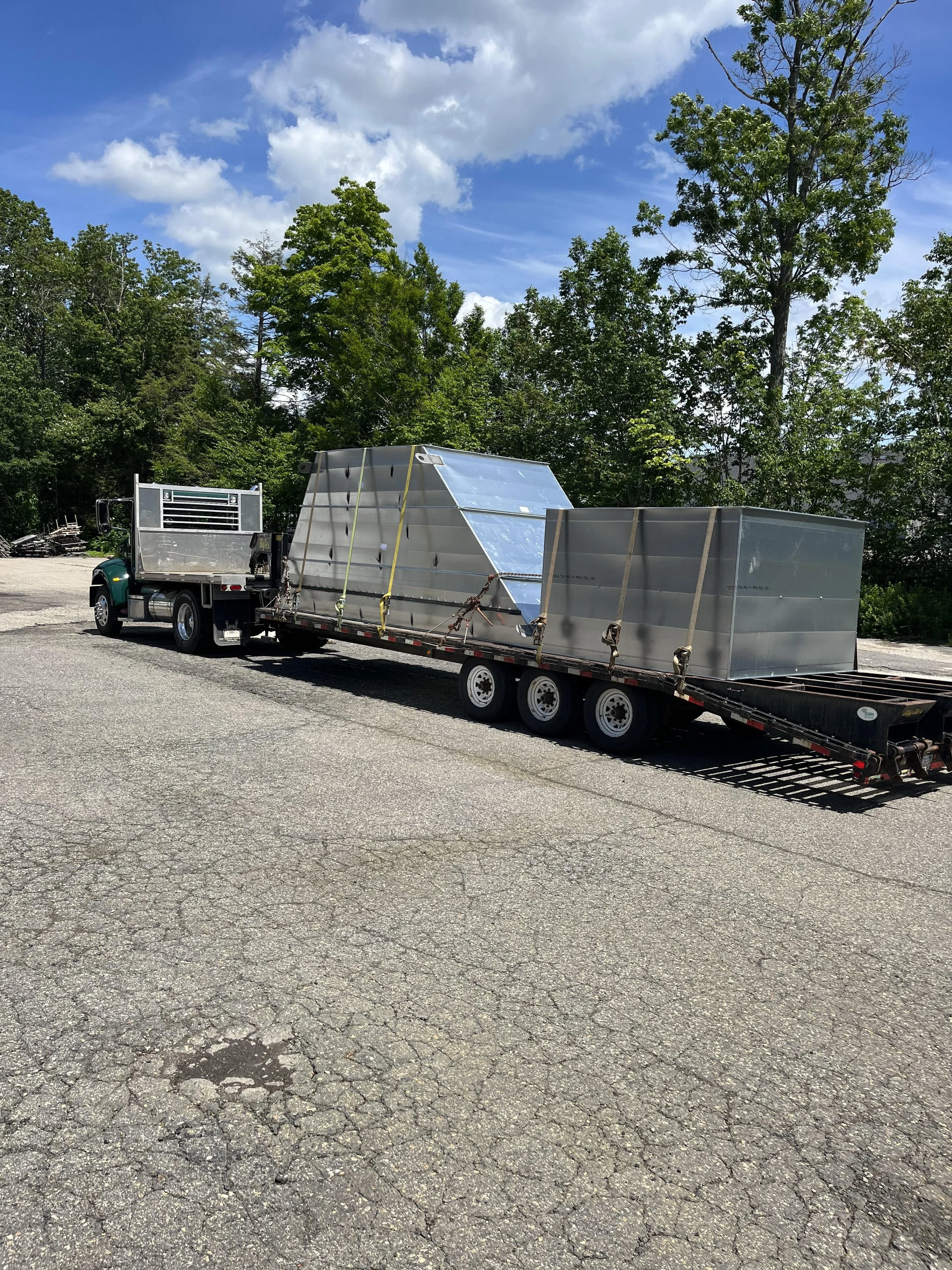 A flatbed truck parked on a paved surface with large metal structures secured on the trailer, surrounded by green trees and a partly cloudy sky.