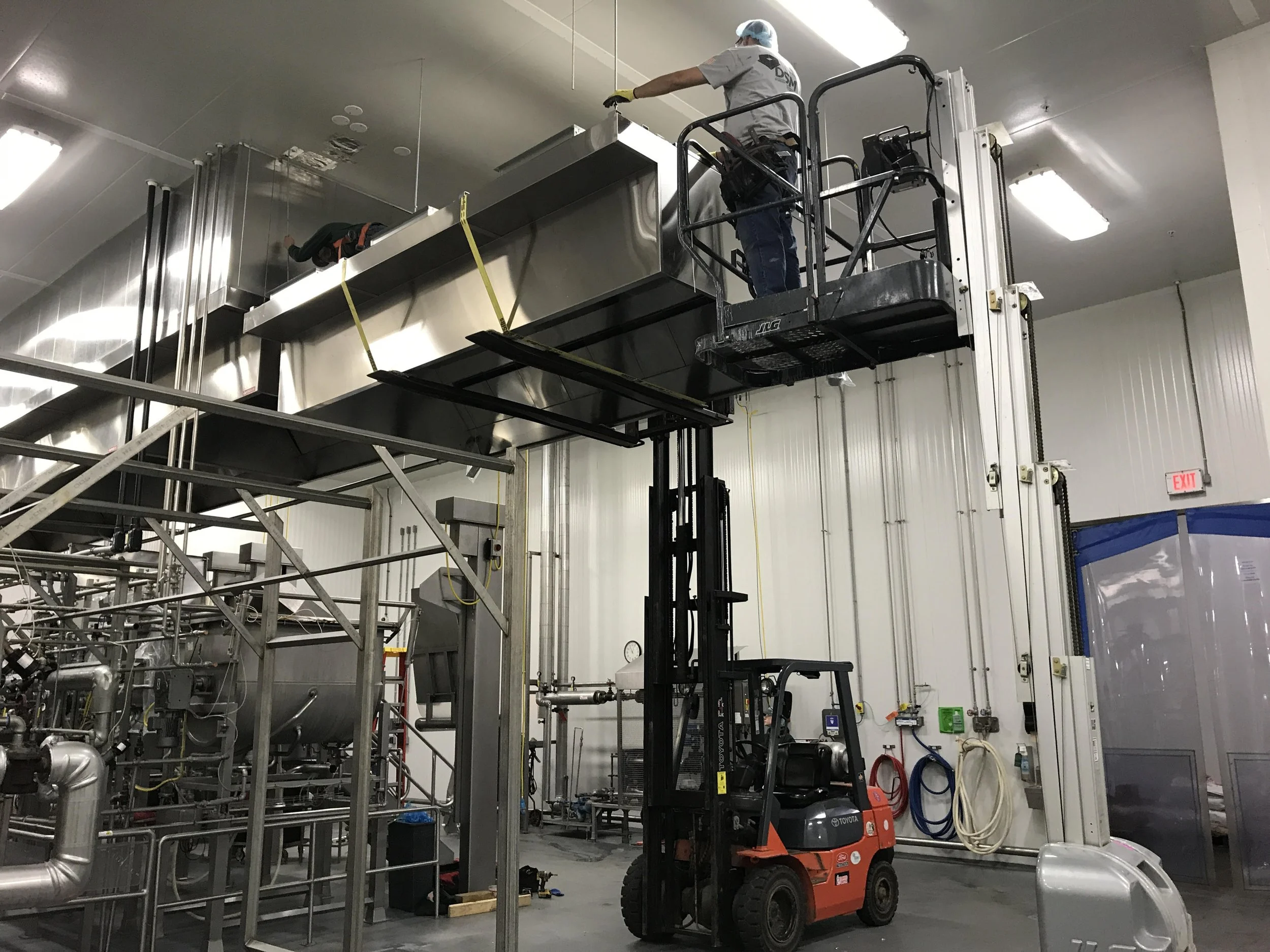 A worker operating a forklift in an industrial setting, lifting a large metal piece near ceiling with piping and industrial equipment in the background.
