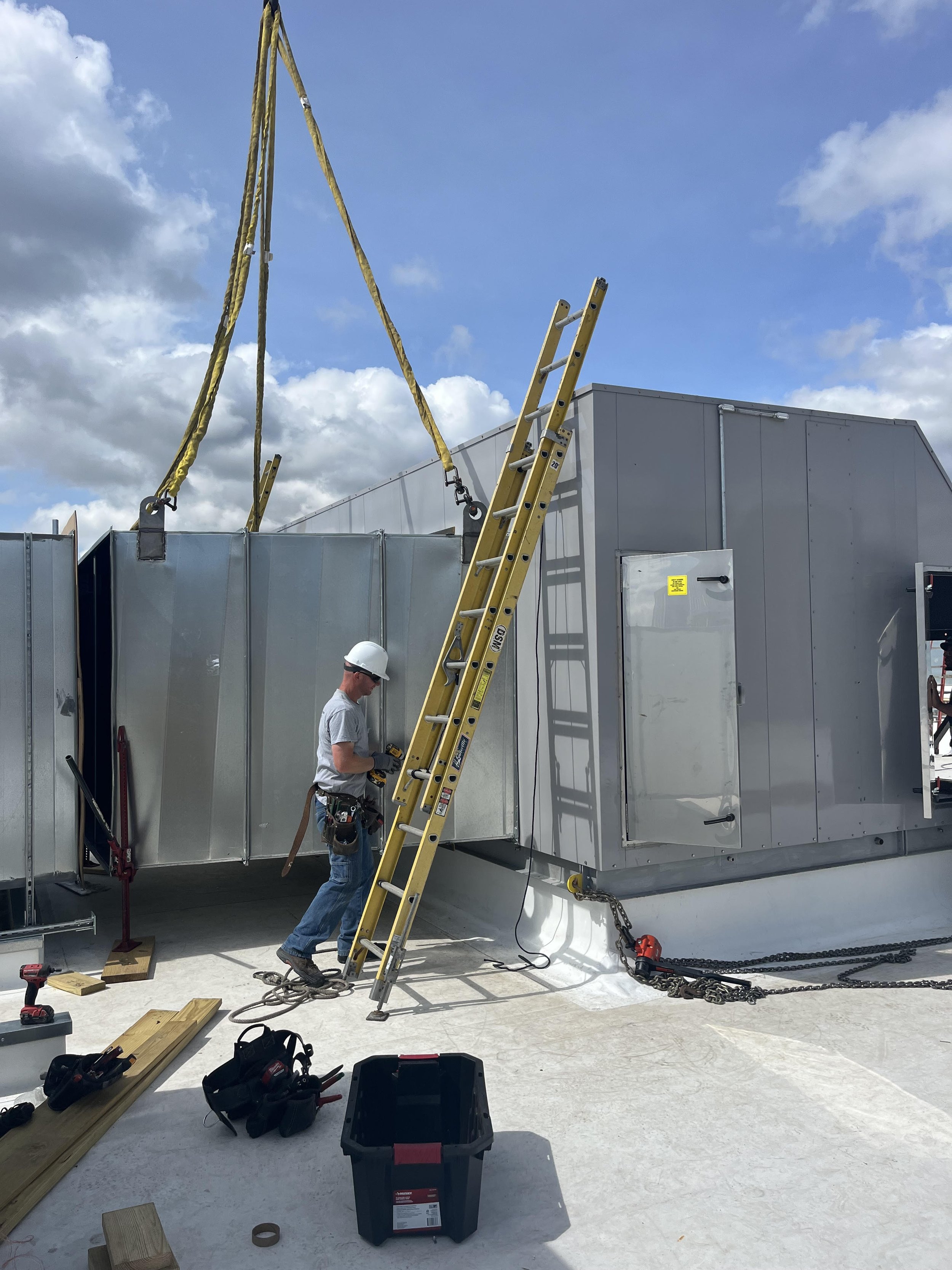 Construction workers installing HVAC duct system, with ladders, tools, and equipment outdoors under a blue sky with clouds.
